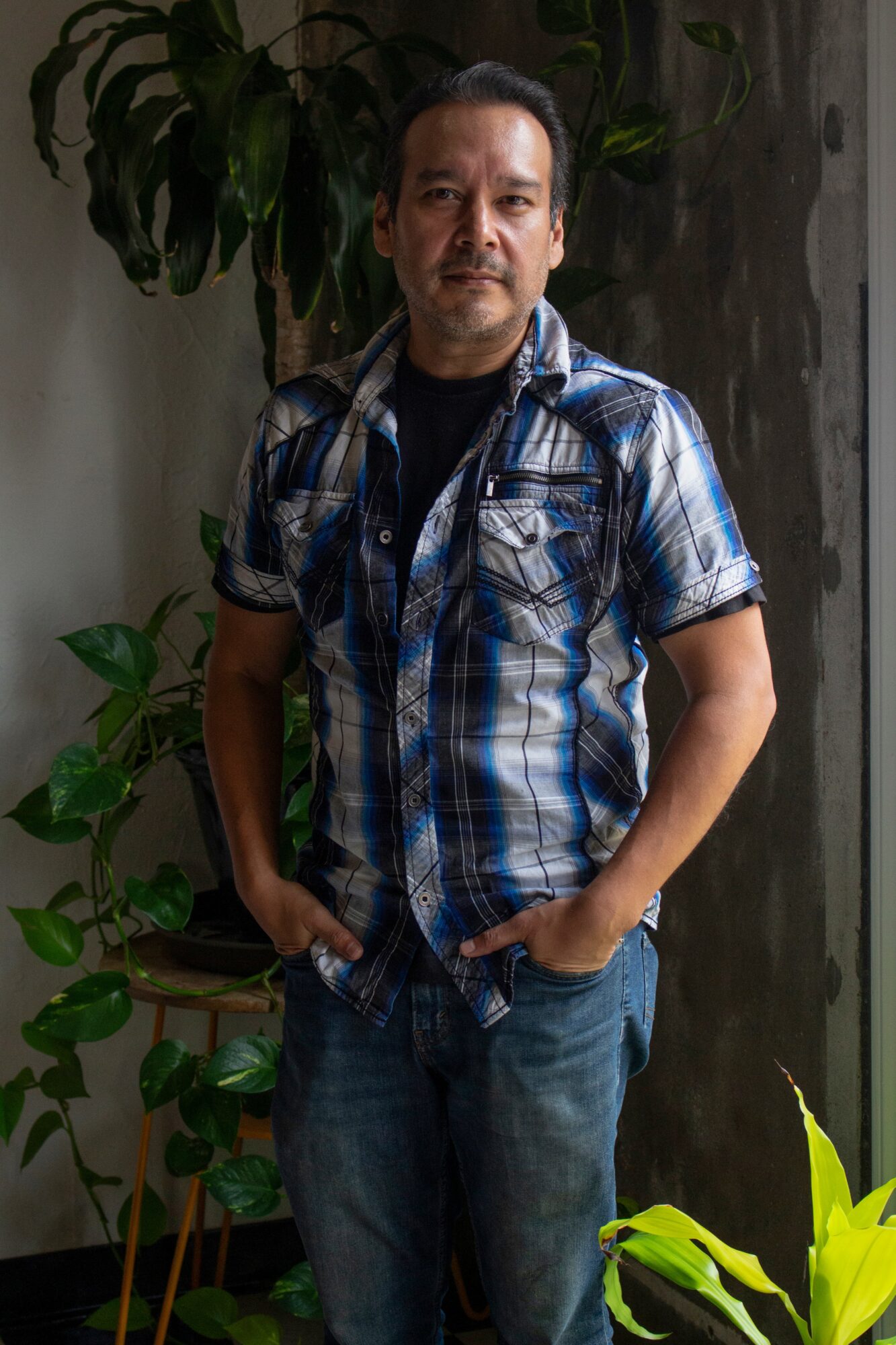 Man with short dark hair and mustache wearing a blue plaid shirt and jeans, standing indoors near plants.