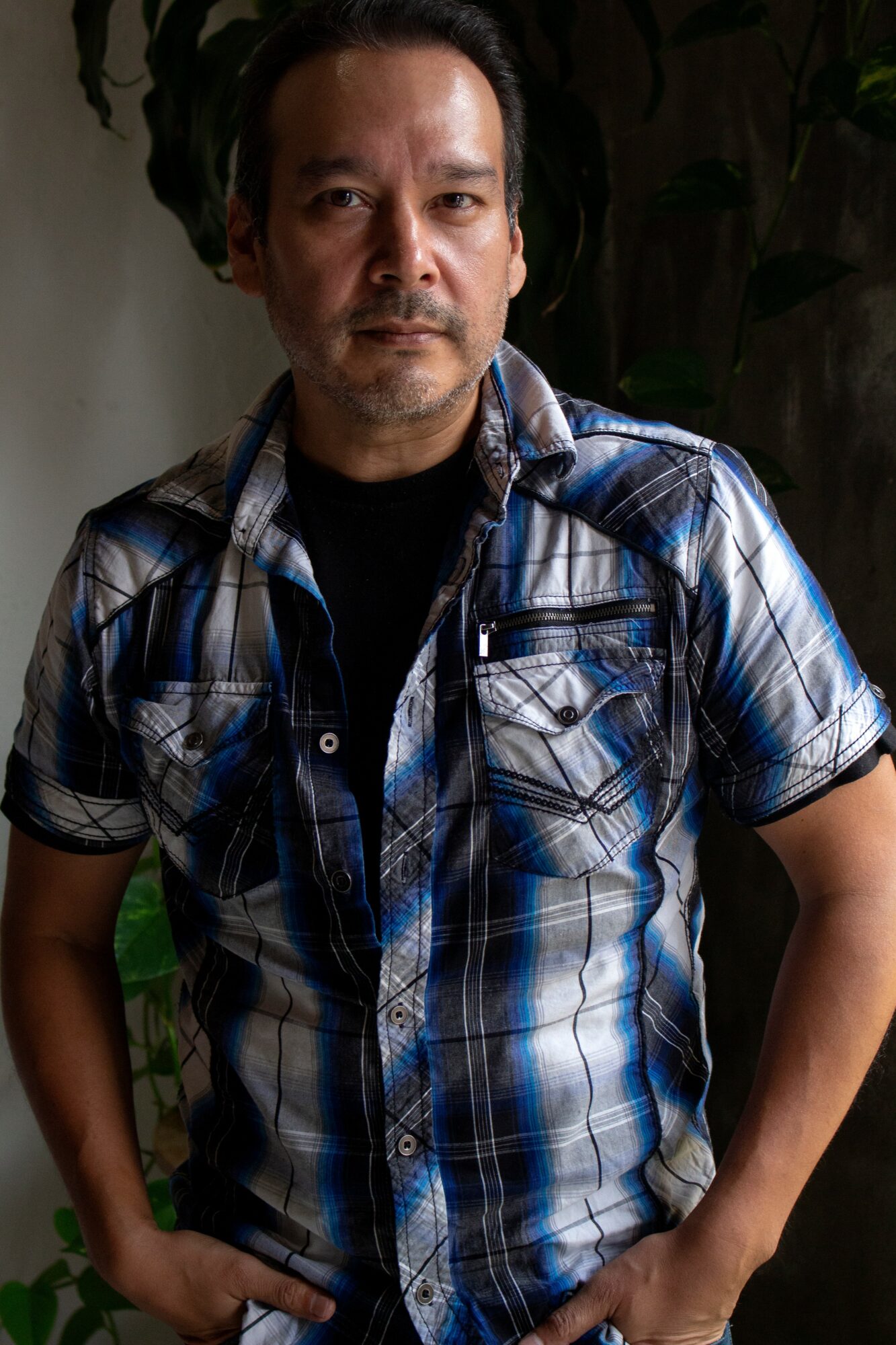 Man with short dark hair wearing a blue plaid shirt, standing outdoors with plants in background.
