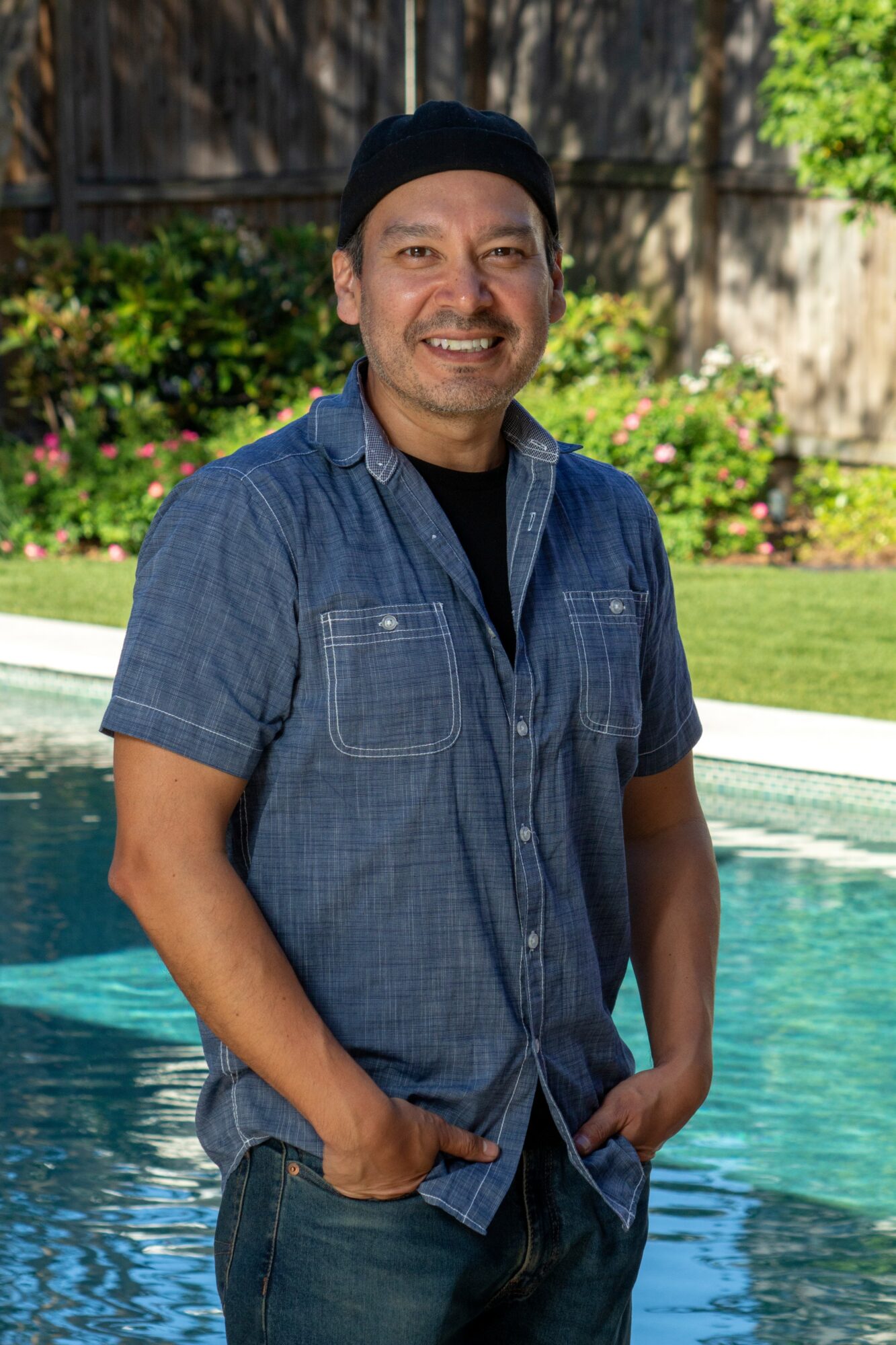 Man smiling outdoors near a pool, wearing a black beanie and denim shirt, with greenery in the background.