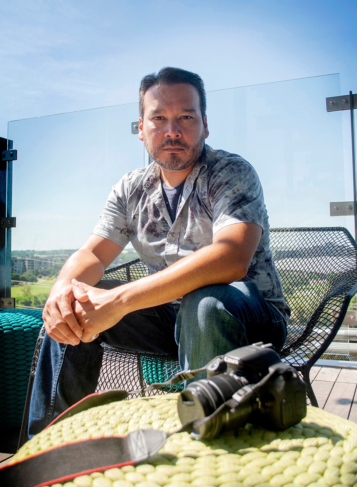 Man with short dark hair and beard sitting outdoors on a metal chair, with a clear glass barrier behind him, holding binoculars on a yellow surface.