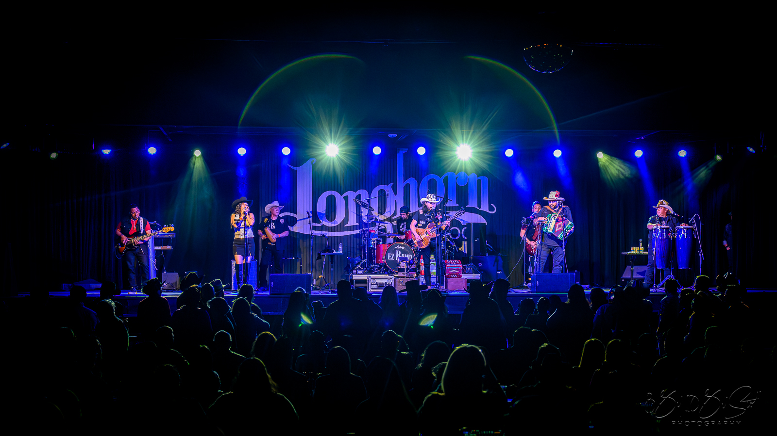 Band performs on stage with blue and green lighting, audience silhouettes visible, and a large banner reading 'Lorchorn' in the background.