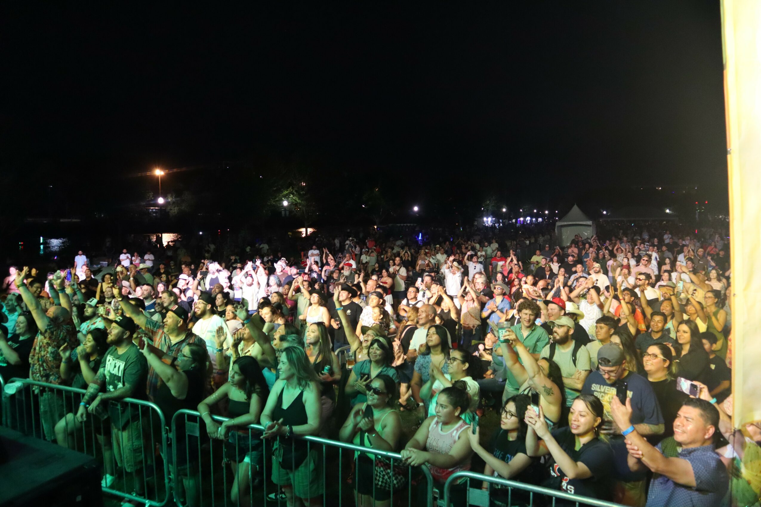 Crowd of people at an outdoor event during nighttime, some raising hands, with barriers in front, illuminated by stage lights.