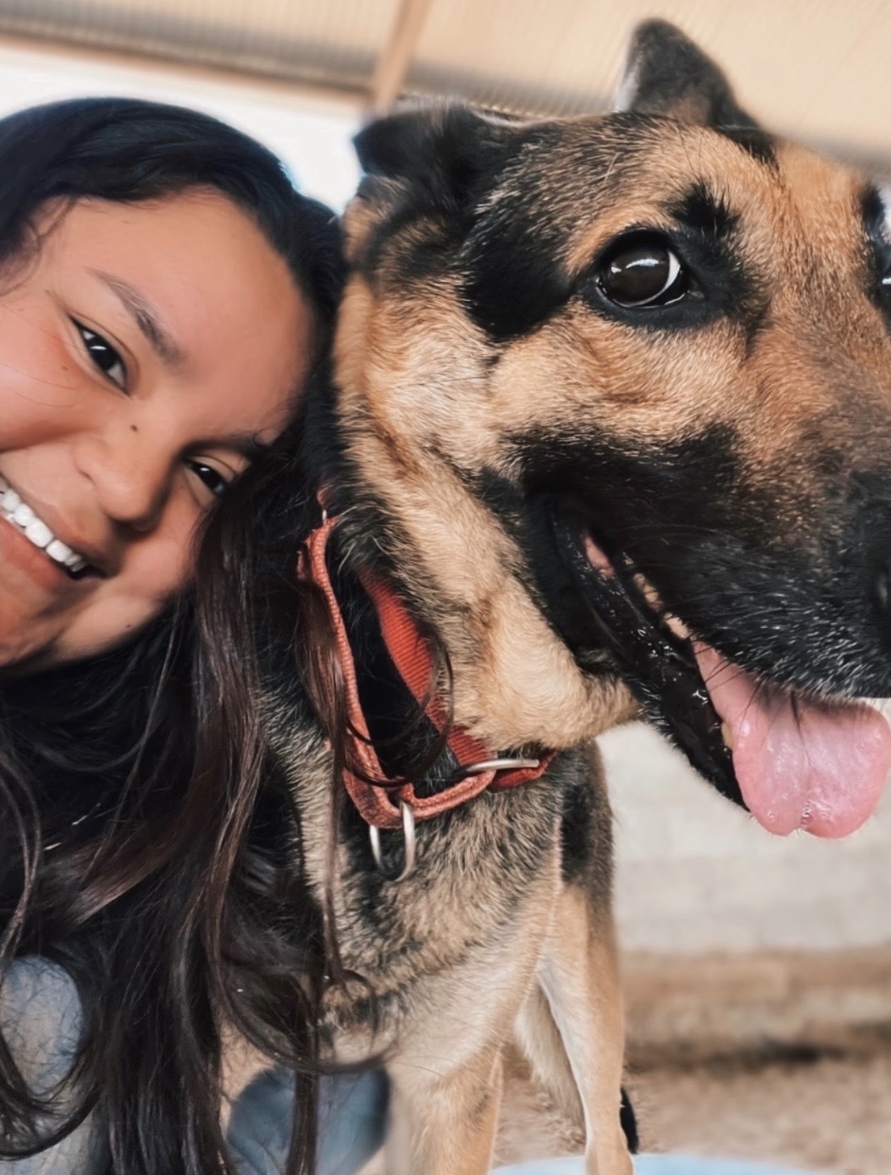 Person with long dark hair smiling next to a large dog with a black and tan coat, open mouth, and tongue out.
