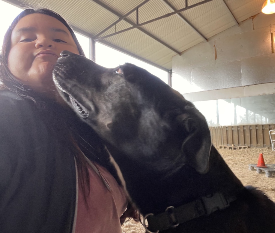 Person with a black dog inside a building, the dog appears to be licking the person's face.