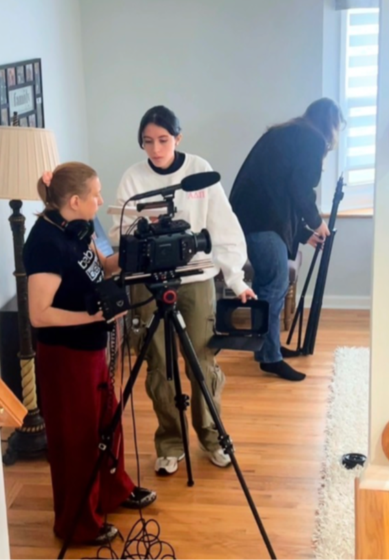 Three people in a room with a camera on a tripod, two women and a girl, preparing for filming, wooden floor, window with blinds.