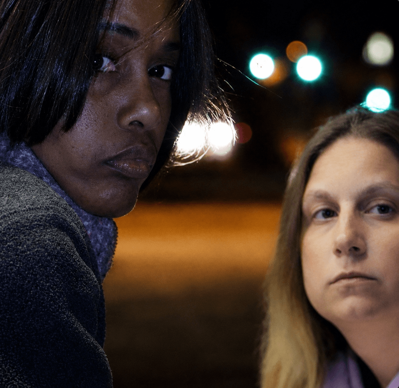 Two women with dark and light hair, close-up, nighttime with blurred city lights in background.