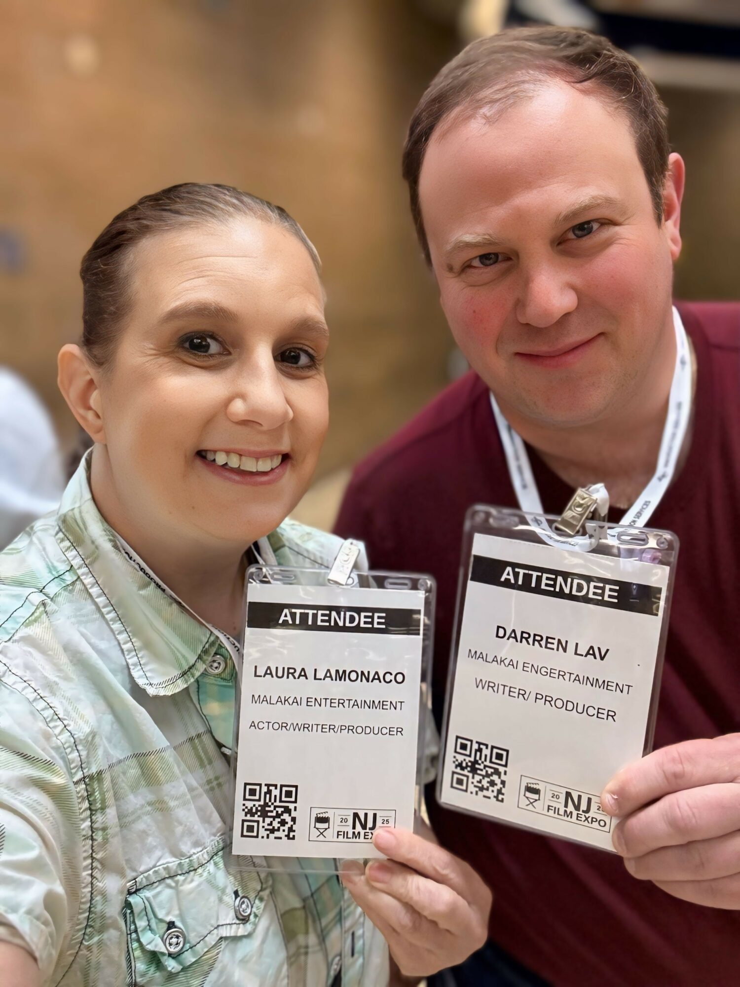 Two people holding conference badges, smiling, with a blurred background, one woman and one man, both looking at the camera.