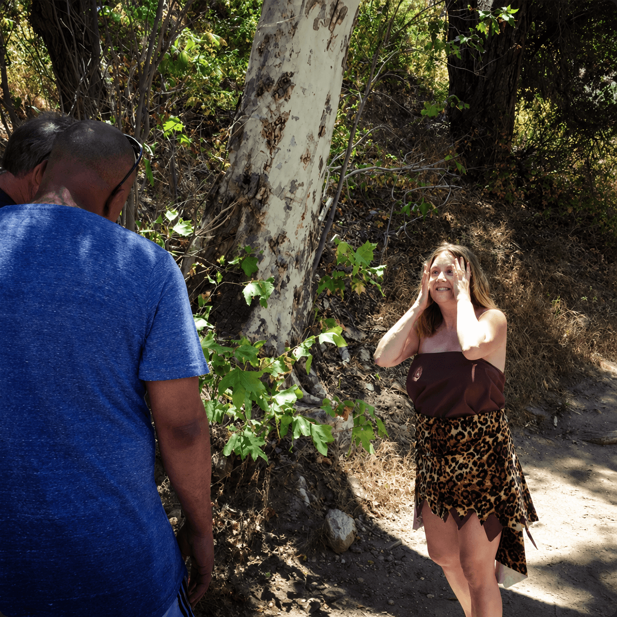 Woman with long hair and leopard print skirt smiling outdoors, talking to a person in a blue shirt, trees in background.