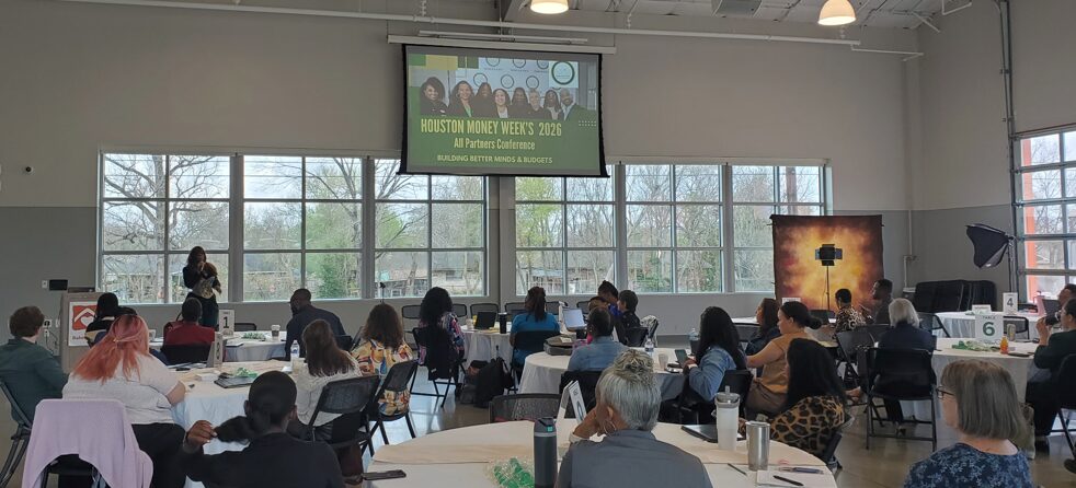 Conference room with people seated at tables, large windows, and a screen displaying a presentation.