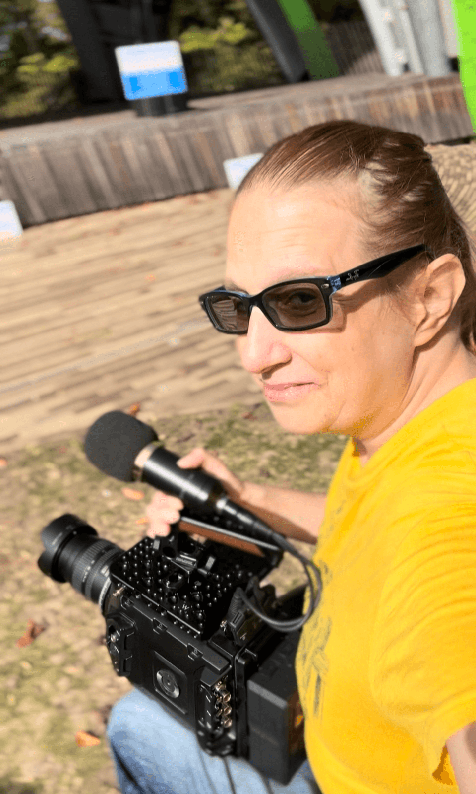 Woman wearing sunglasses and a yellow shirt holding a camera outdoors.