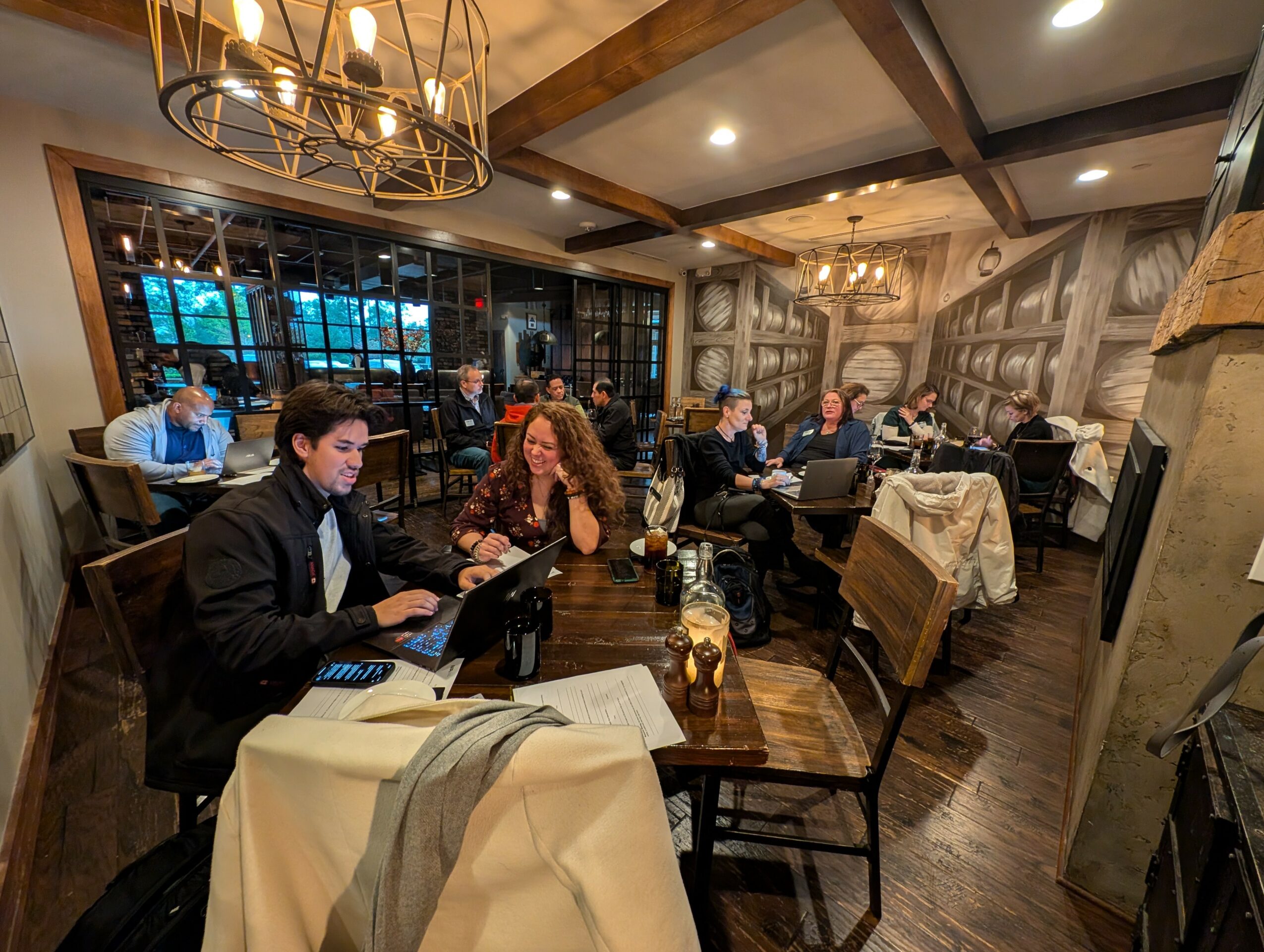 People dining in a cozy restaurant with wooden beams, large windows, and warm lighting, engaged in conversation and using devices.