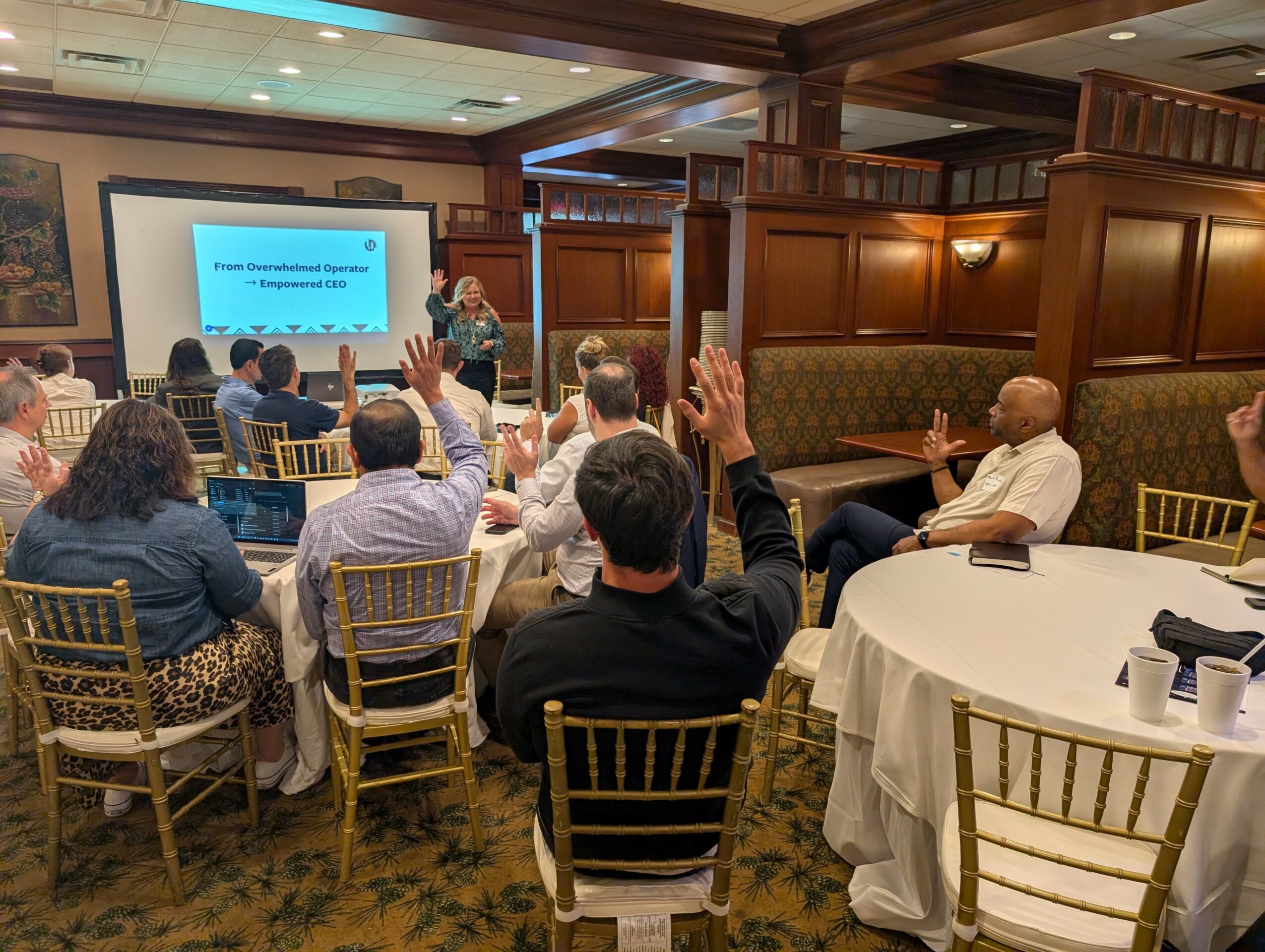 People seated at round tables in a conference room, some raising hands, facing a presenter and screen, wood-paneled walls.