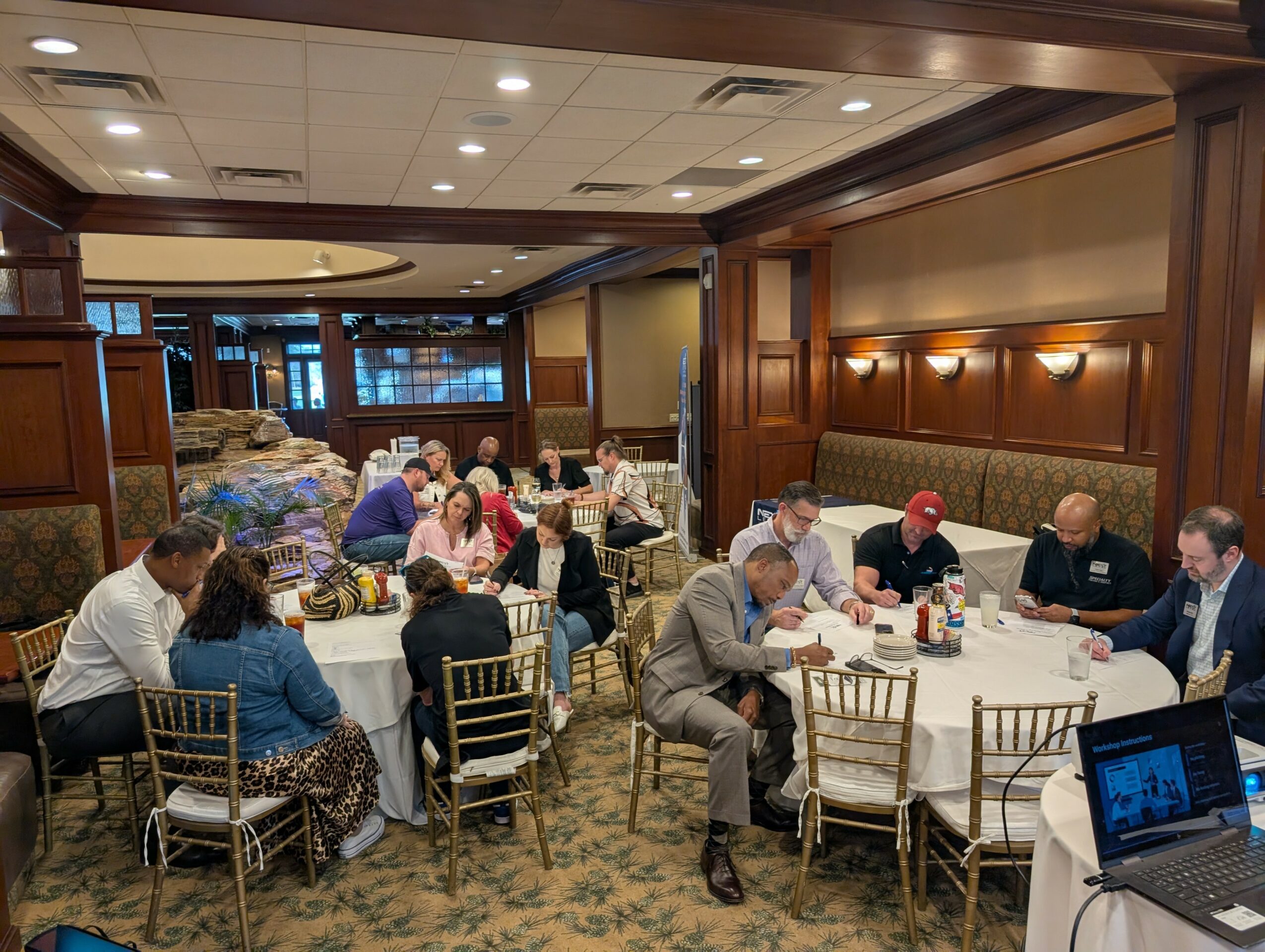 People seated at round tables in a banquet room, engaging in conversation and dining, with wood-paneled walls and warm lighting.