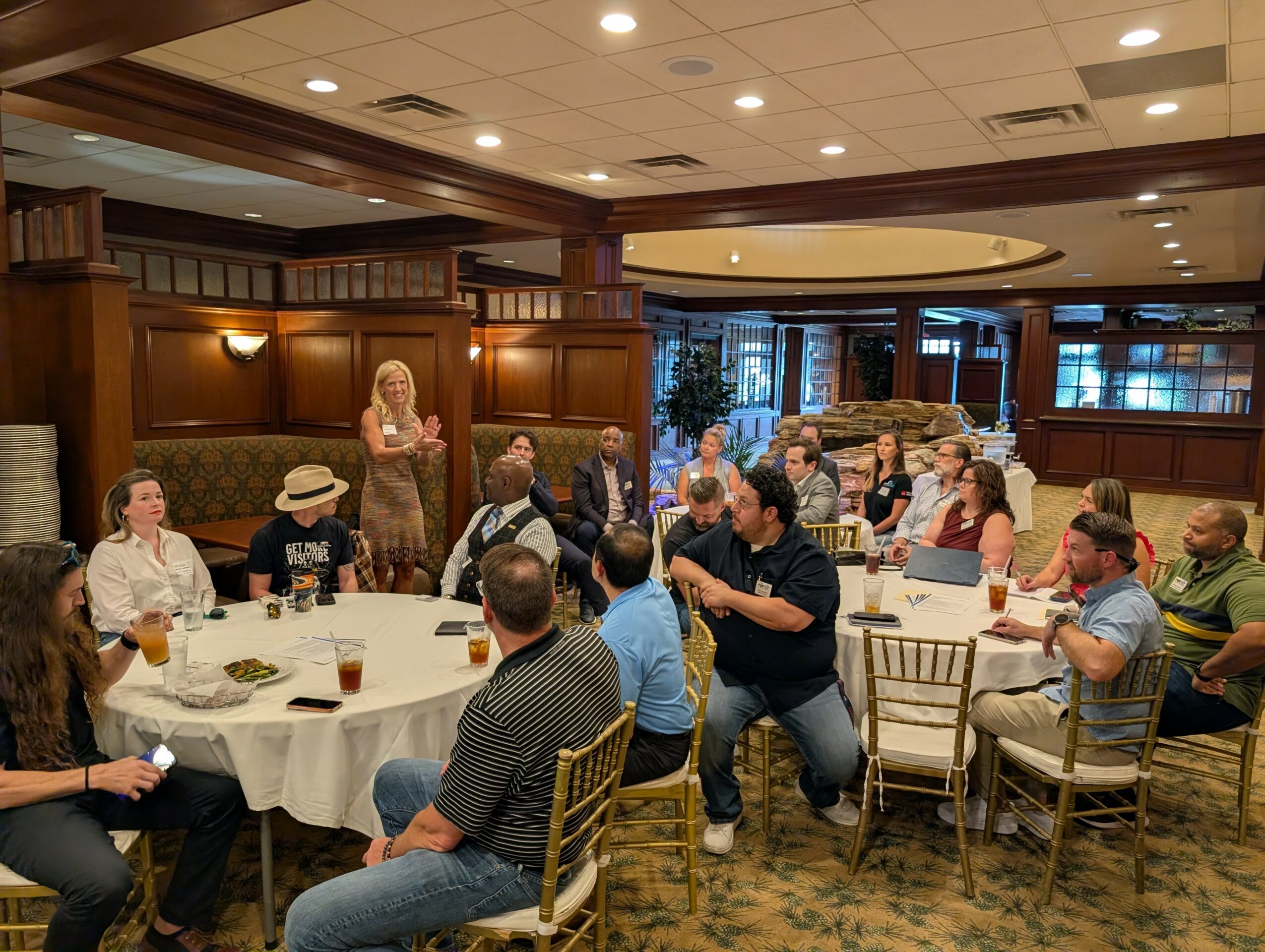 People seated around a round table in a restaurant, some standing and talking, with a woman speaking at the front.
