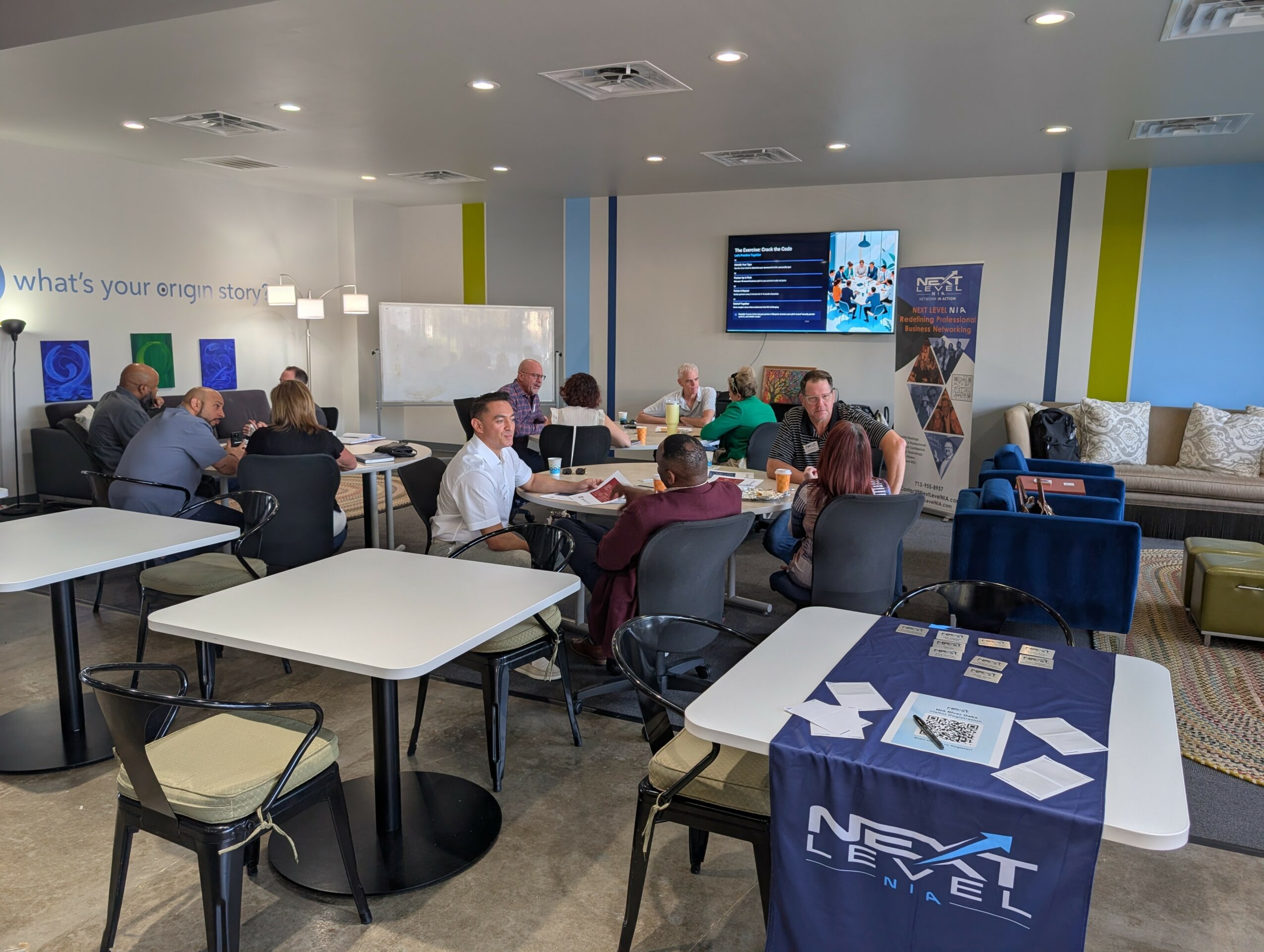 People seated at tables in a conference room with a large screen and whiteboard, some working on laptops, others talking.