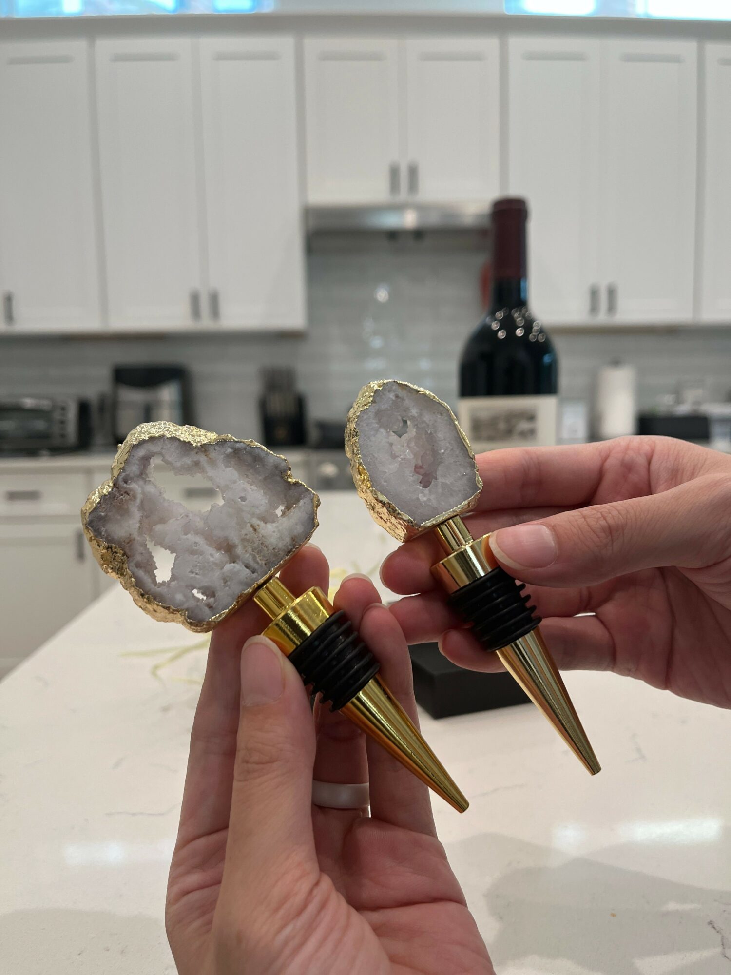 Hands holding two wine stoppers with large geode crystals, gold and black accents, in a kitchen setting.