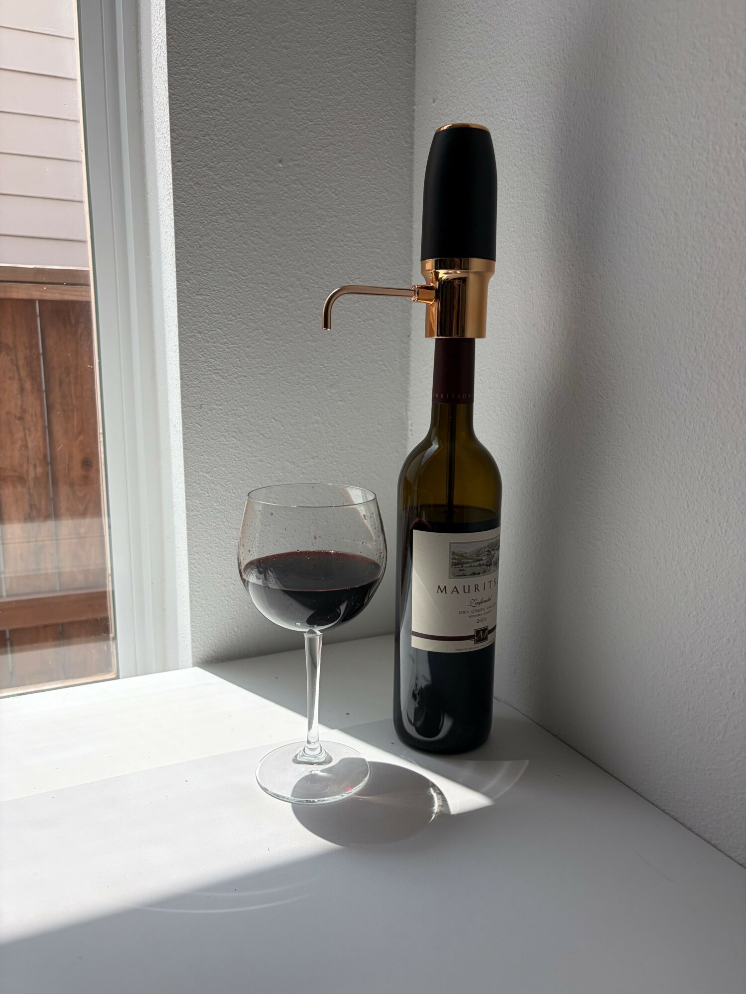 Wine bottle resting on a wall, with a glass of red wine on a white surface, against a plain wall.