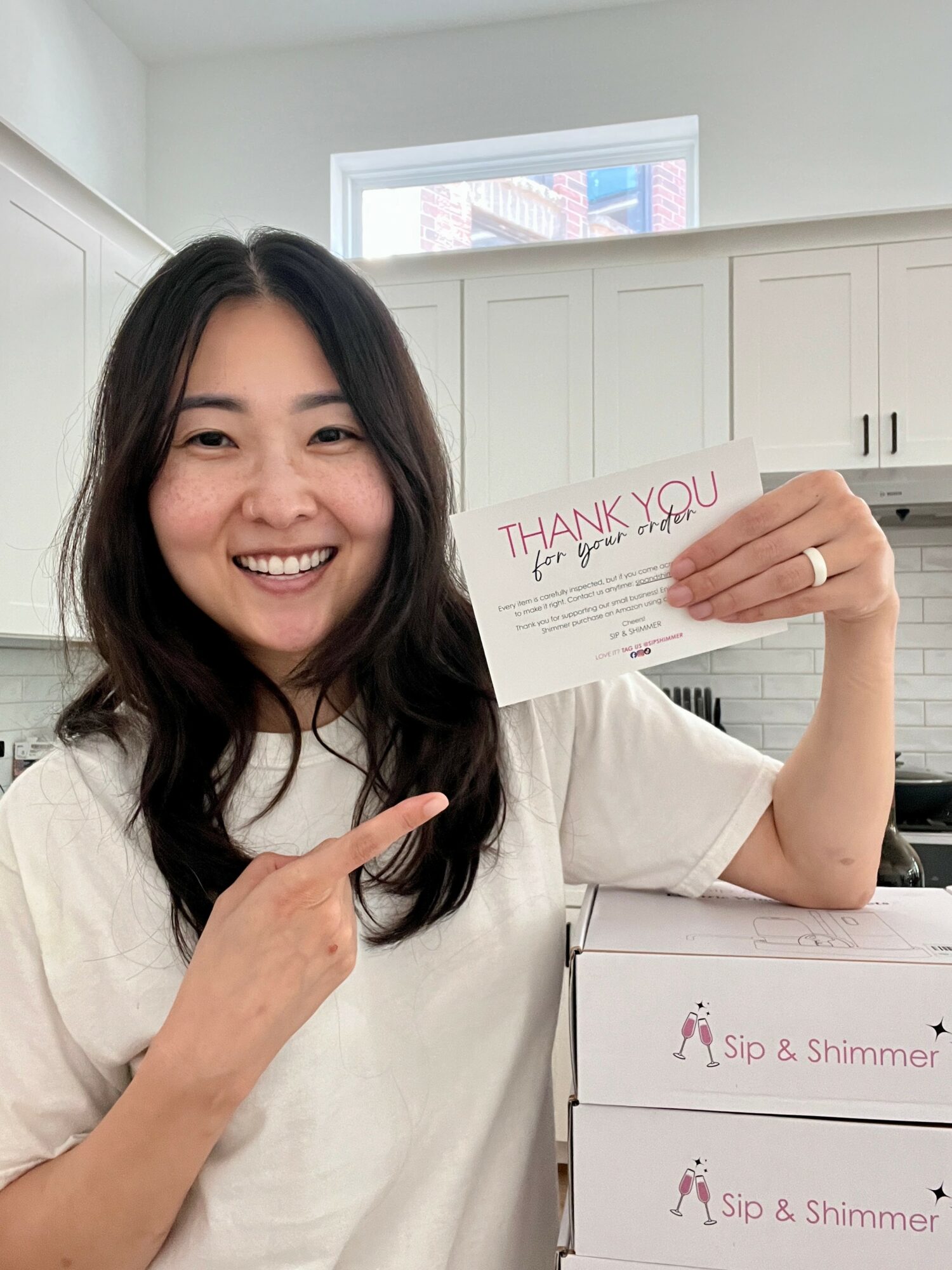 Woman smiling and holding a thank you card, pointing at it, in a bright kitchen.