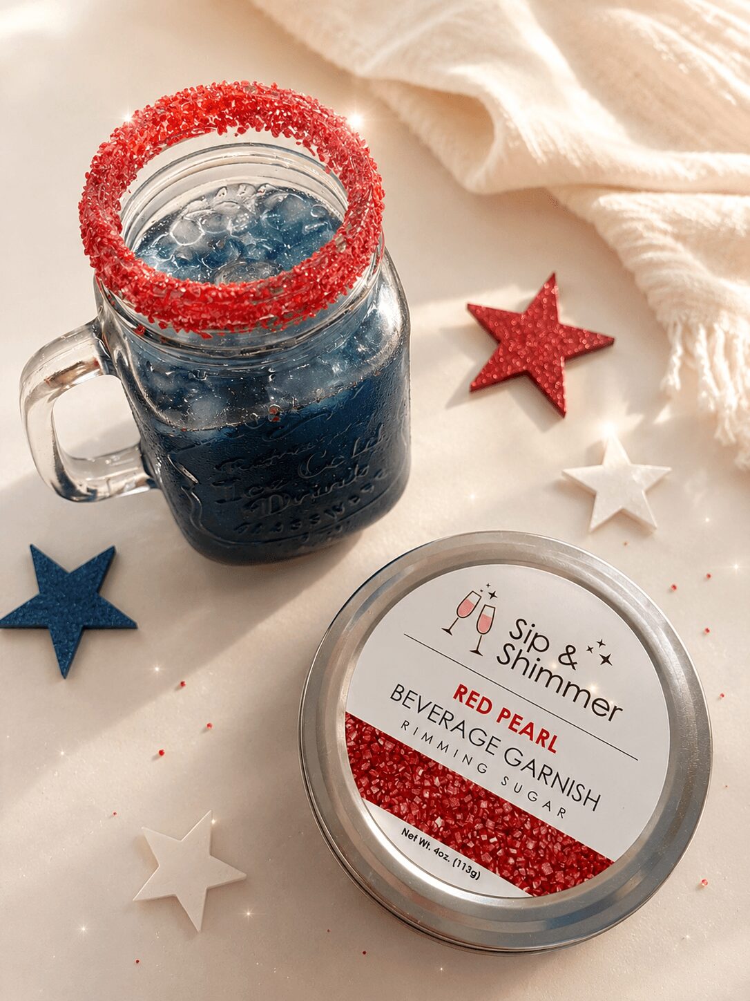 Glass mug filled with dark beverage, decorated with red sugar rim, surrounded by red, white, and blue star-shaped decorations, and a container of red pearl beverage garnish.