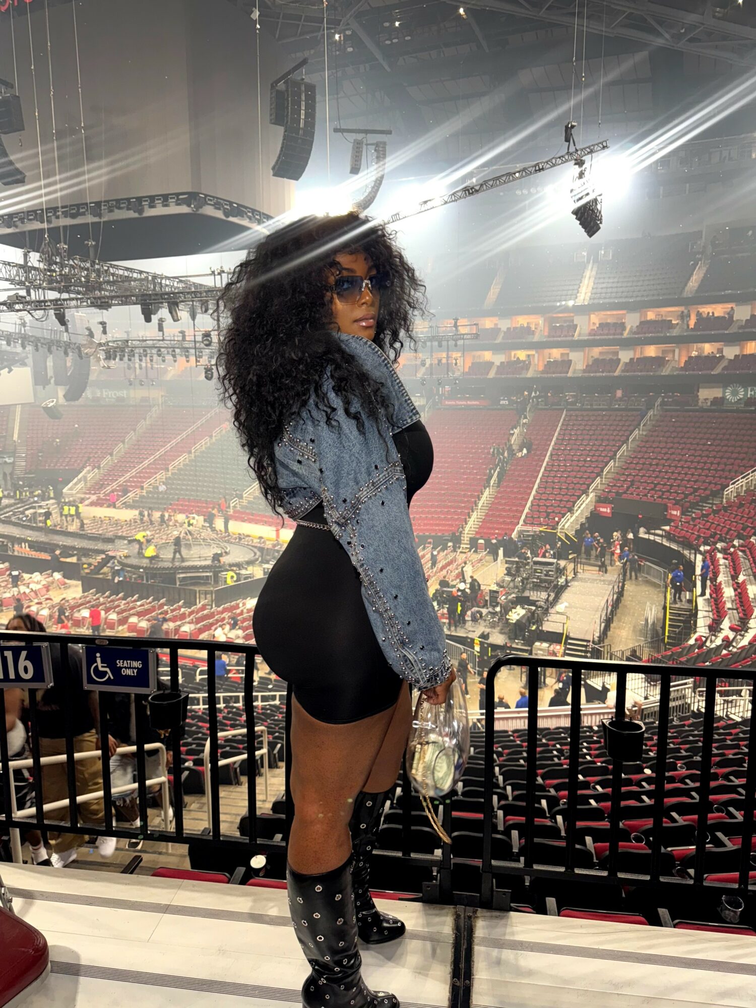 Woman with curly hair and sunglasses standing in an arena, holding a bag, with stage and seating in background.