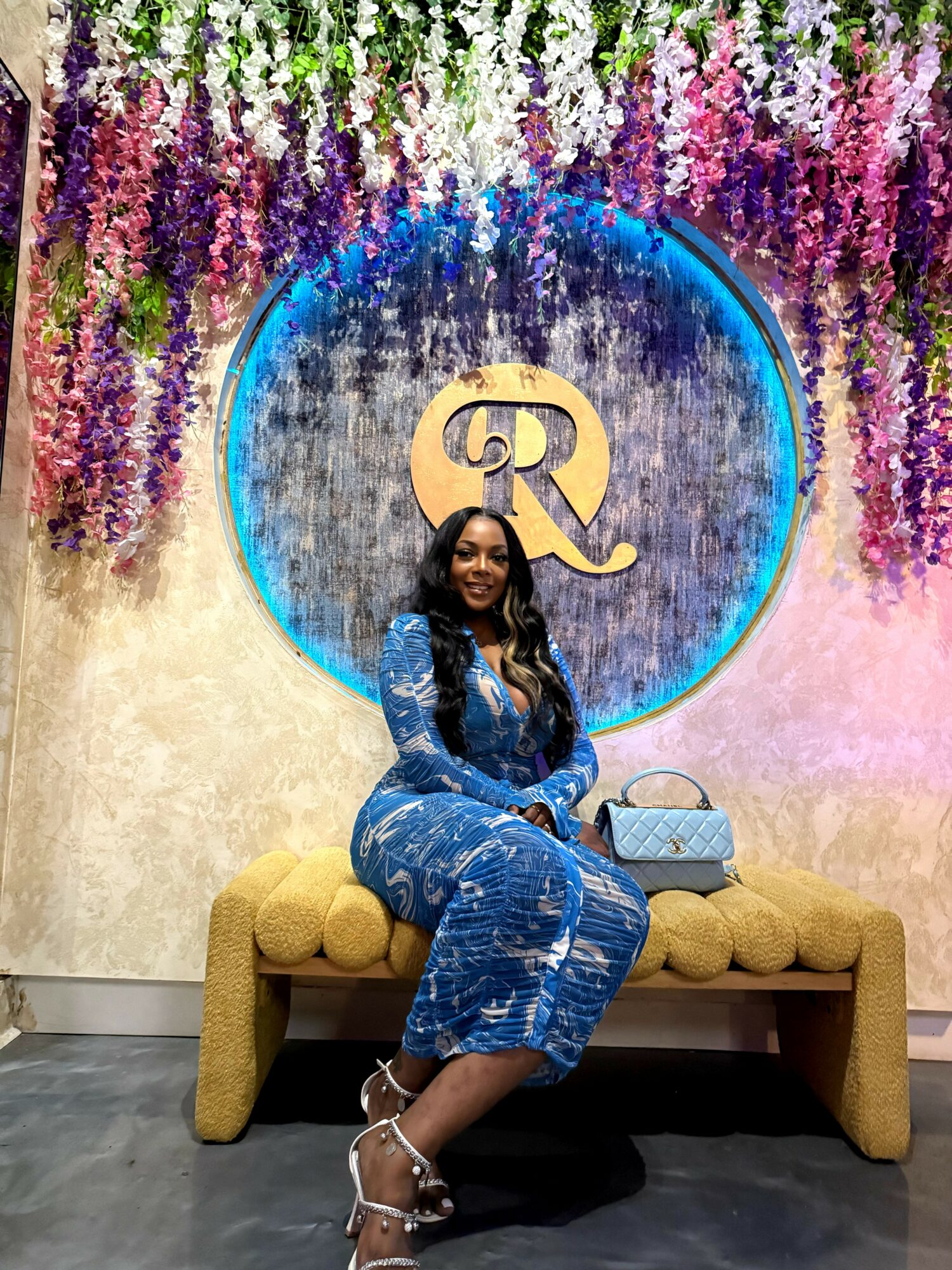 Woman sitting on a yellow bench in front of a decorative wall with purple flowers and a blue circle with gold initials.