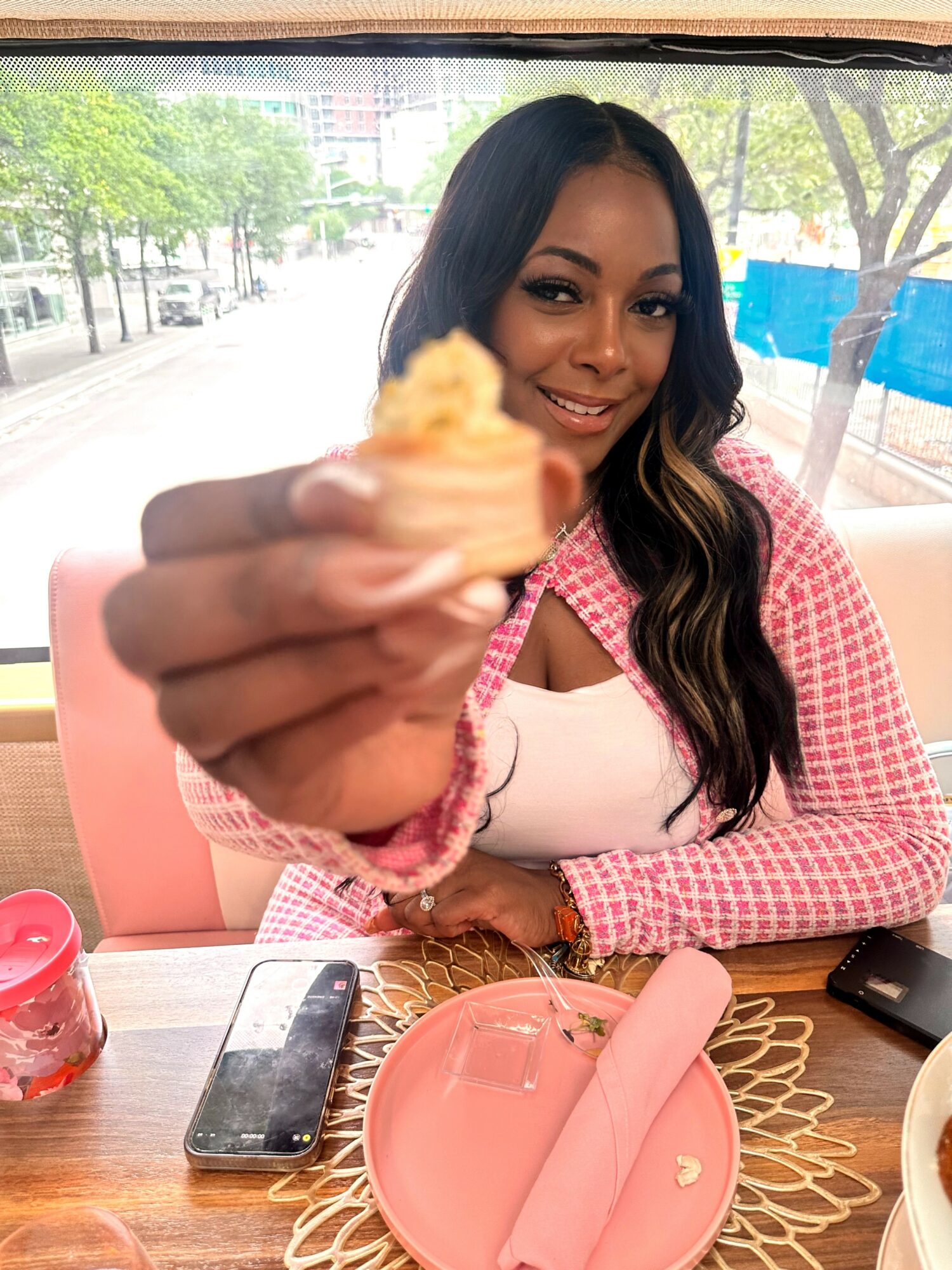 Woman smiling holding a cupcake in a cafe, table with pink plate, napkin, and phone, window with trees outside.