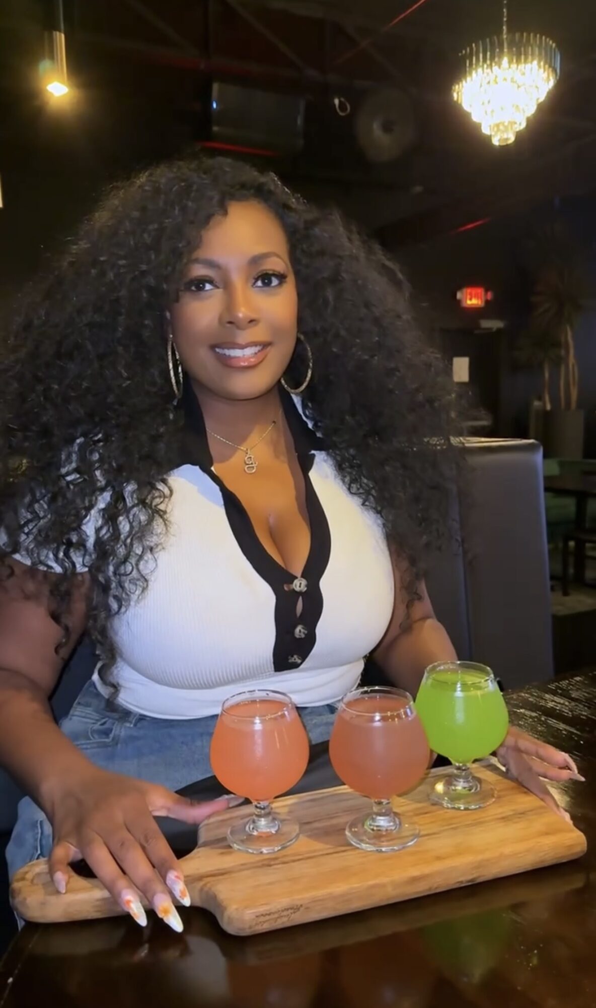 Woman with curly hair smiling at a table with three colorful drinks on a wooden board in a dimly lit restaurant.
