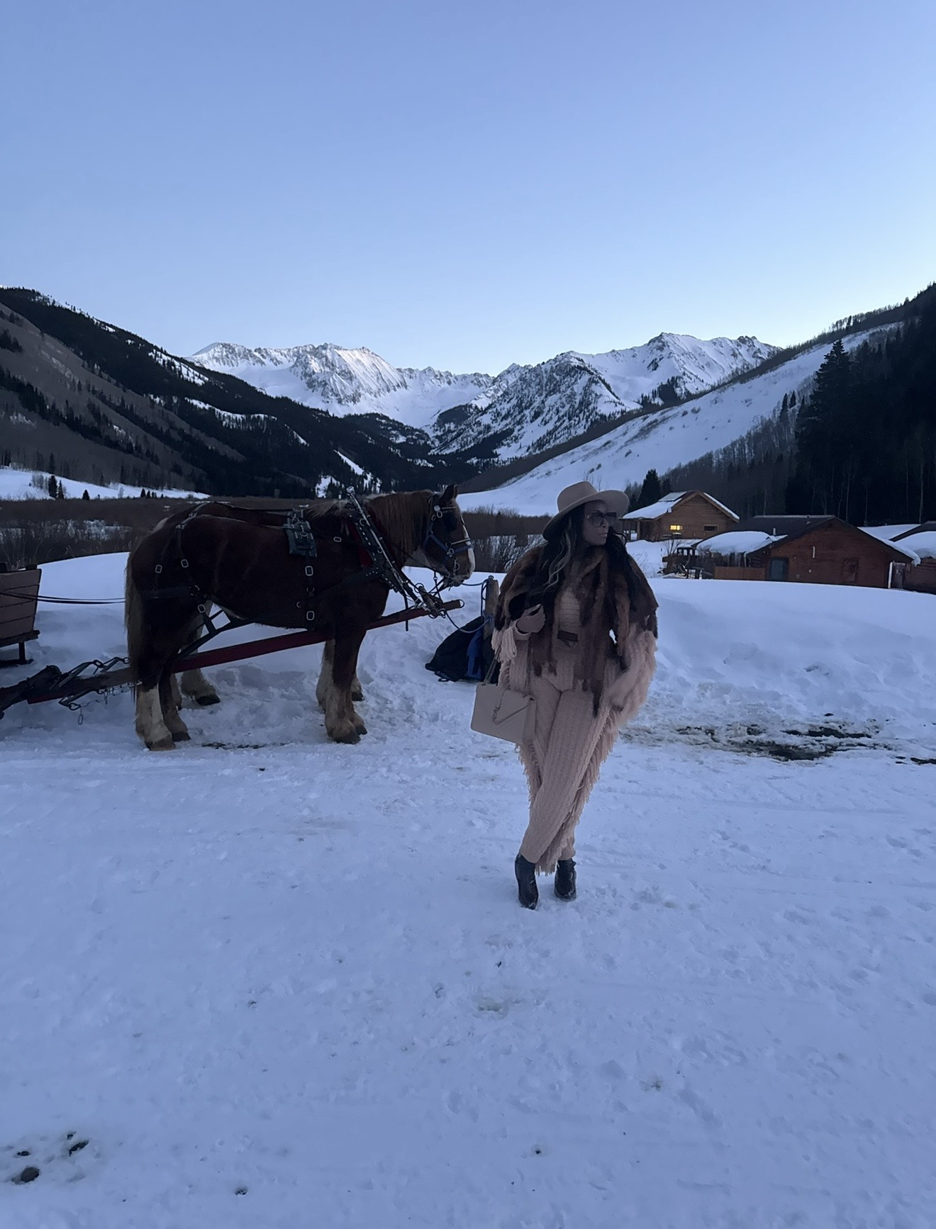 Person walking in snow with horse and mountains in background, clear blue sky, snow-covered landscape, buildings nearby.
