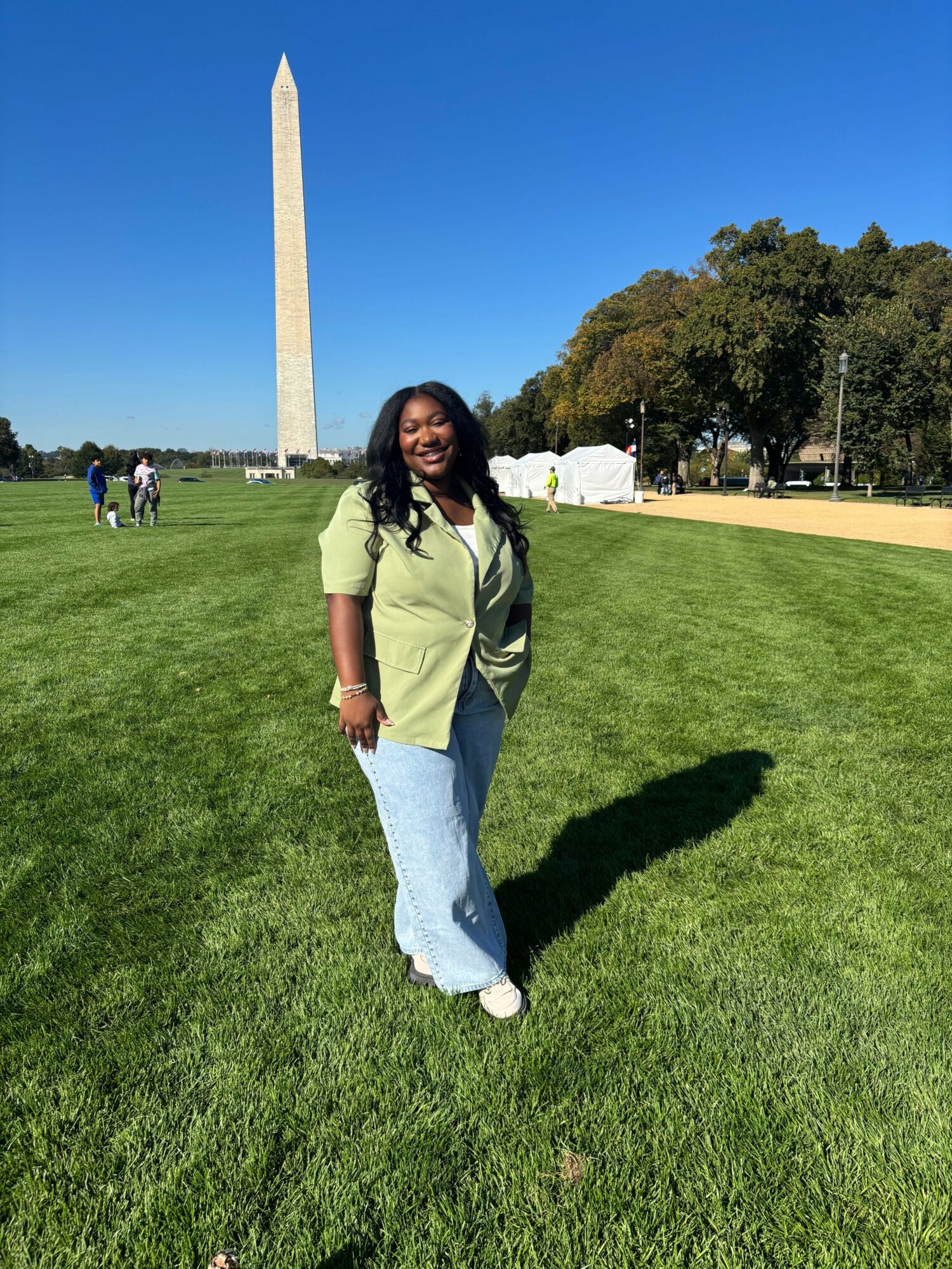 Woman standing on grass near the Washington Monument, with trees and a clear blue sky in the background.