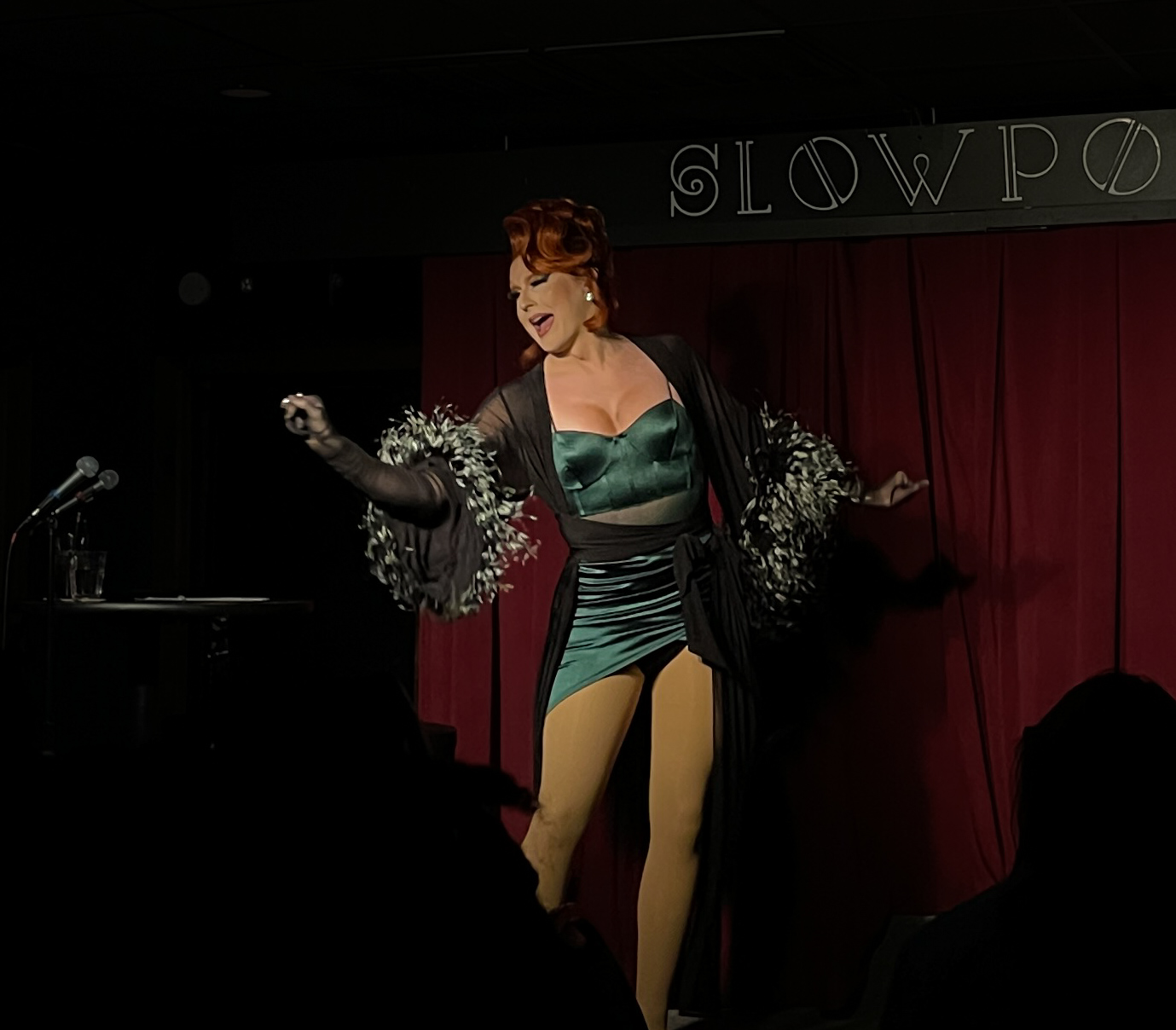 Performer on stage with red curtain, wearing a green dress and black shawl, singing or speaking with arms outstretched.