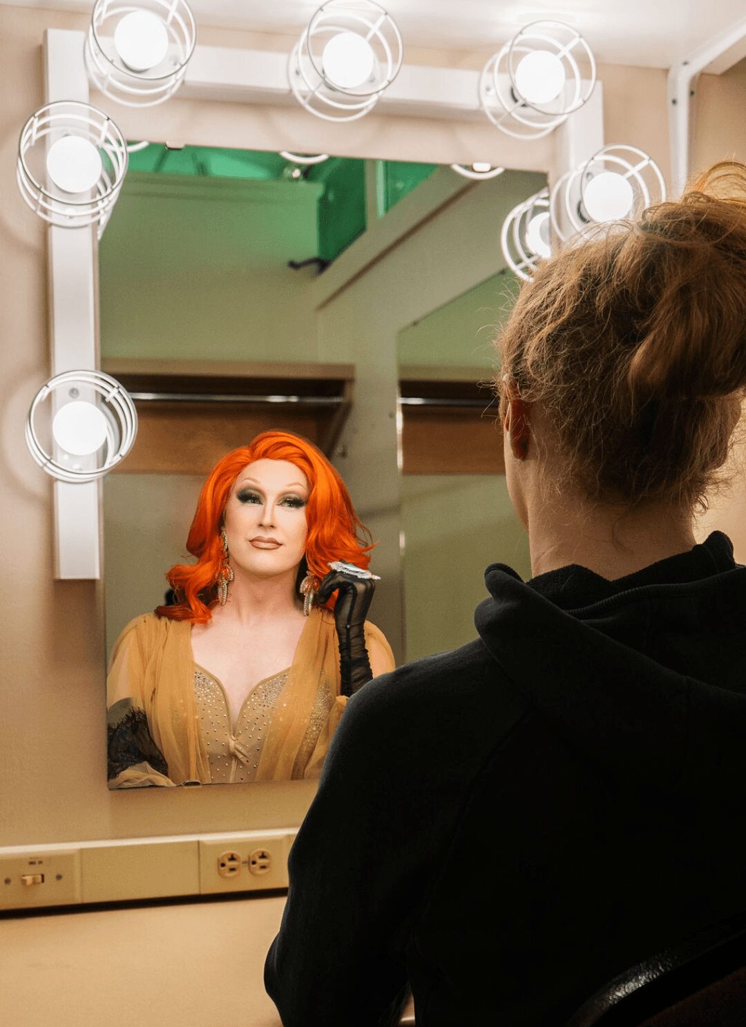 Woman with red hair looking at her reflection in a mirror with bright lights, seen from behind a person with short hair.