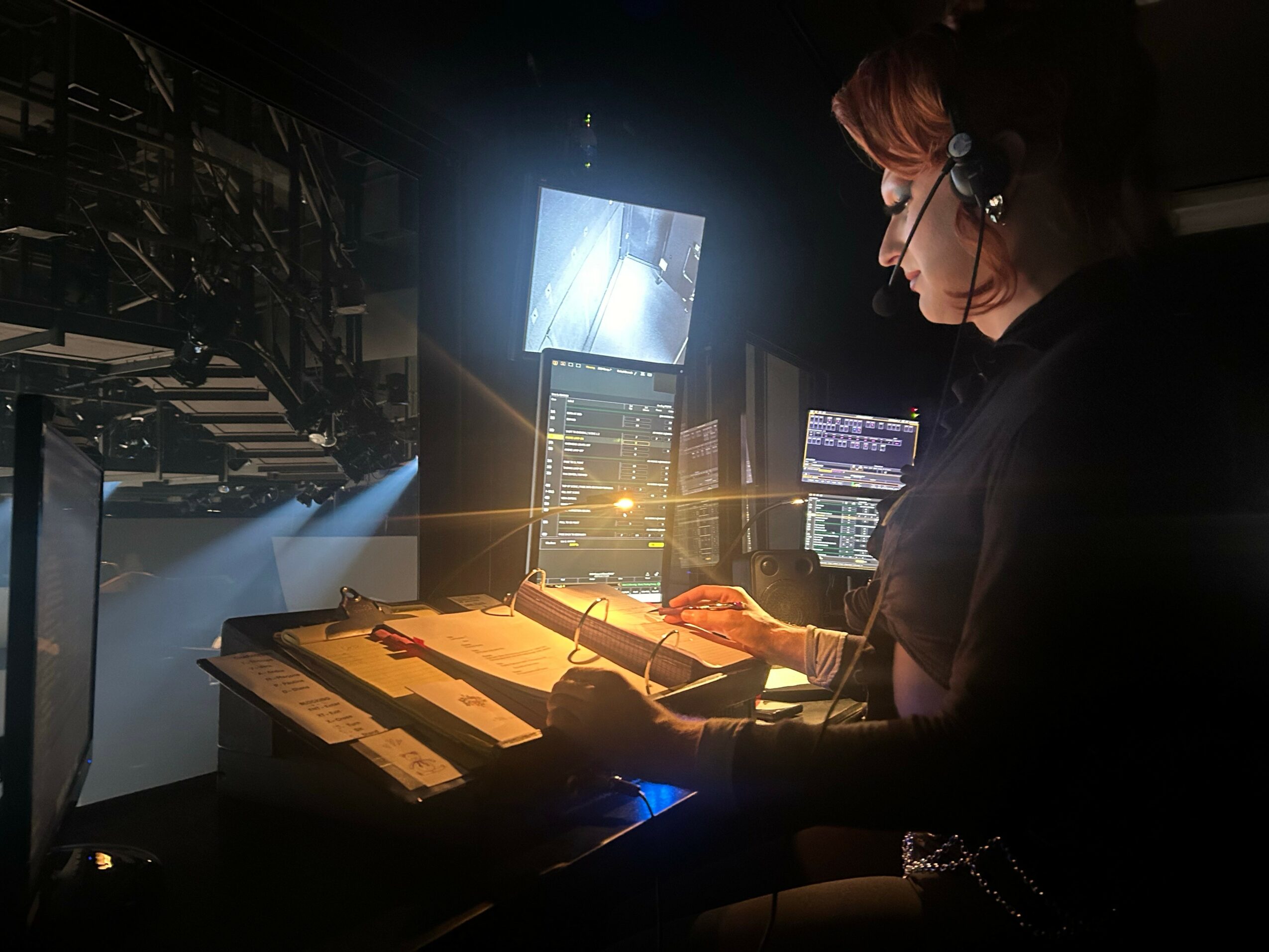 Person wearing headphones working at a control station with multiple screens and equipment in a dark environment.