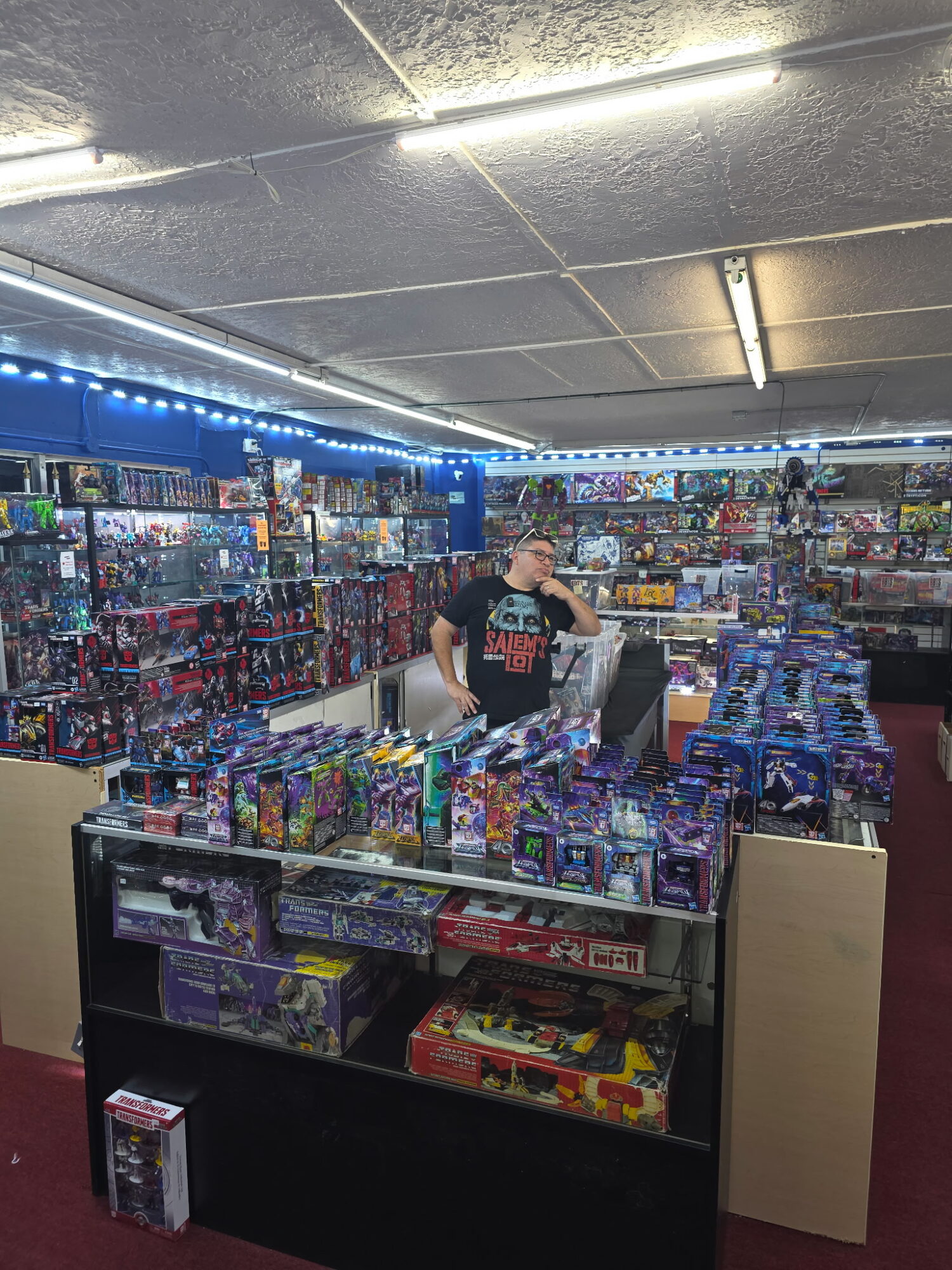 Person standing behind a display of toys in a store aisle, shelves filled with colorful items, ceiling lights overhead.