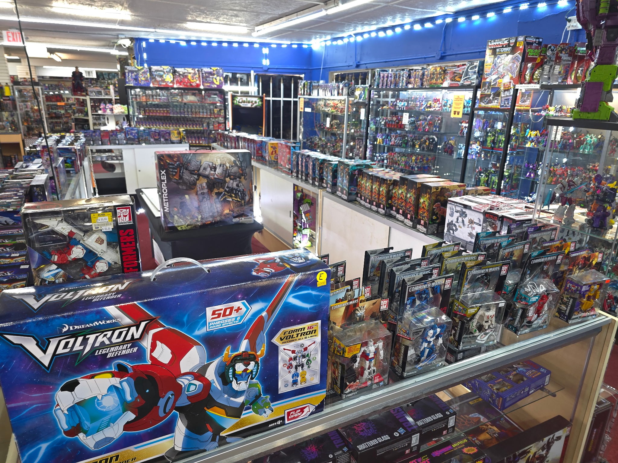 Toy store aisle filled with action figures, model kits, and boxed toys on shelves and display cases, brightly lit.