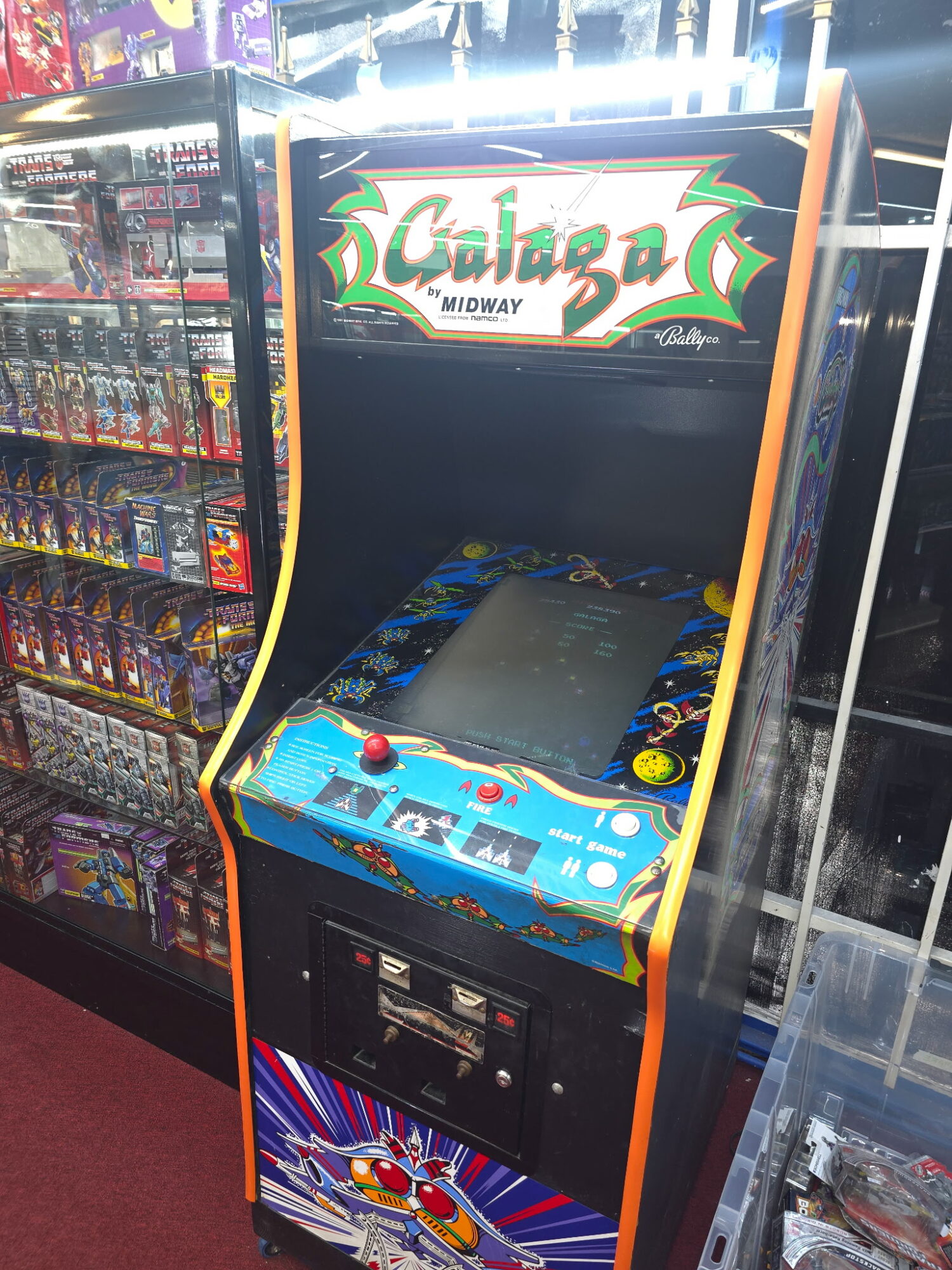 Arcade game machine with colorful design and a control panel, located in a store aisle with snack shelves nearby.