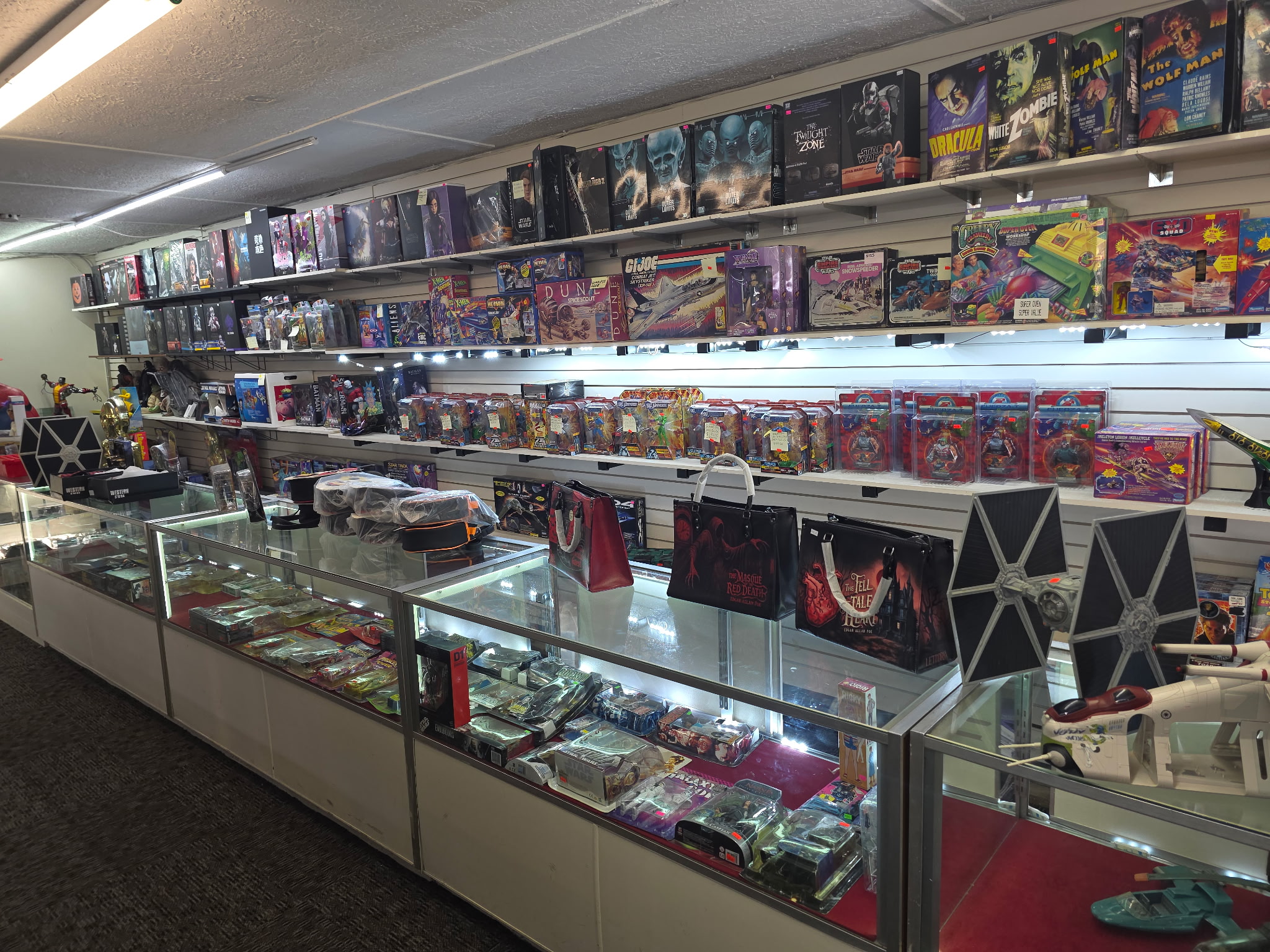 Store display with shelves of board games and collectibles, glass display cases with various items, and a ceiling with fluorescent lights.