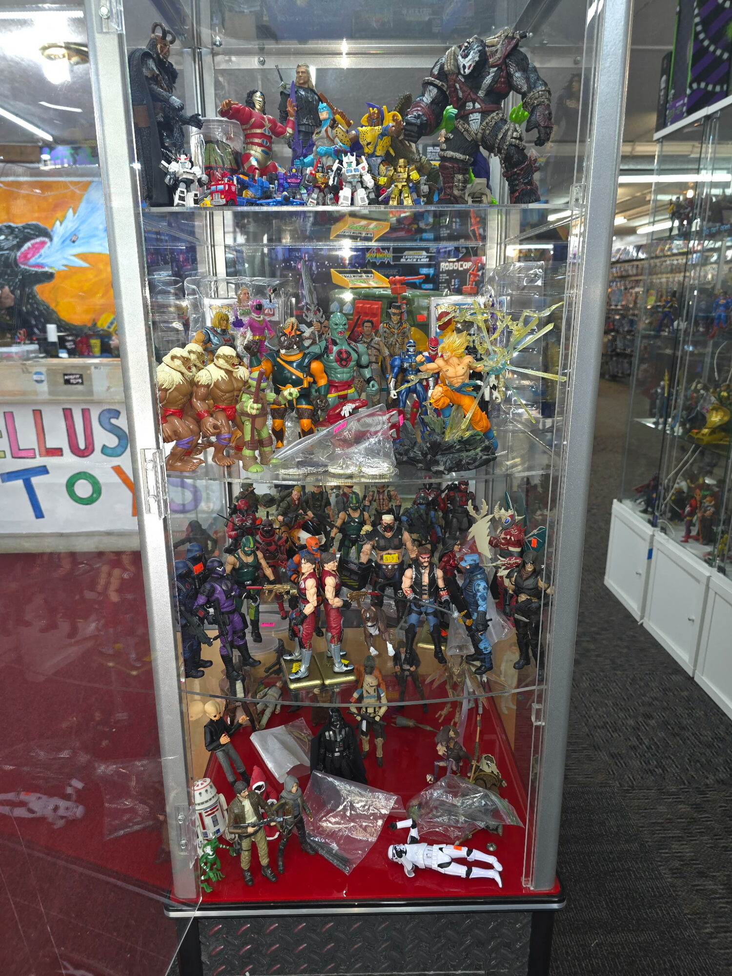 Display case with various action figures and toys arranged on multiple shelves, with reflections and store surroundings visible.
