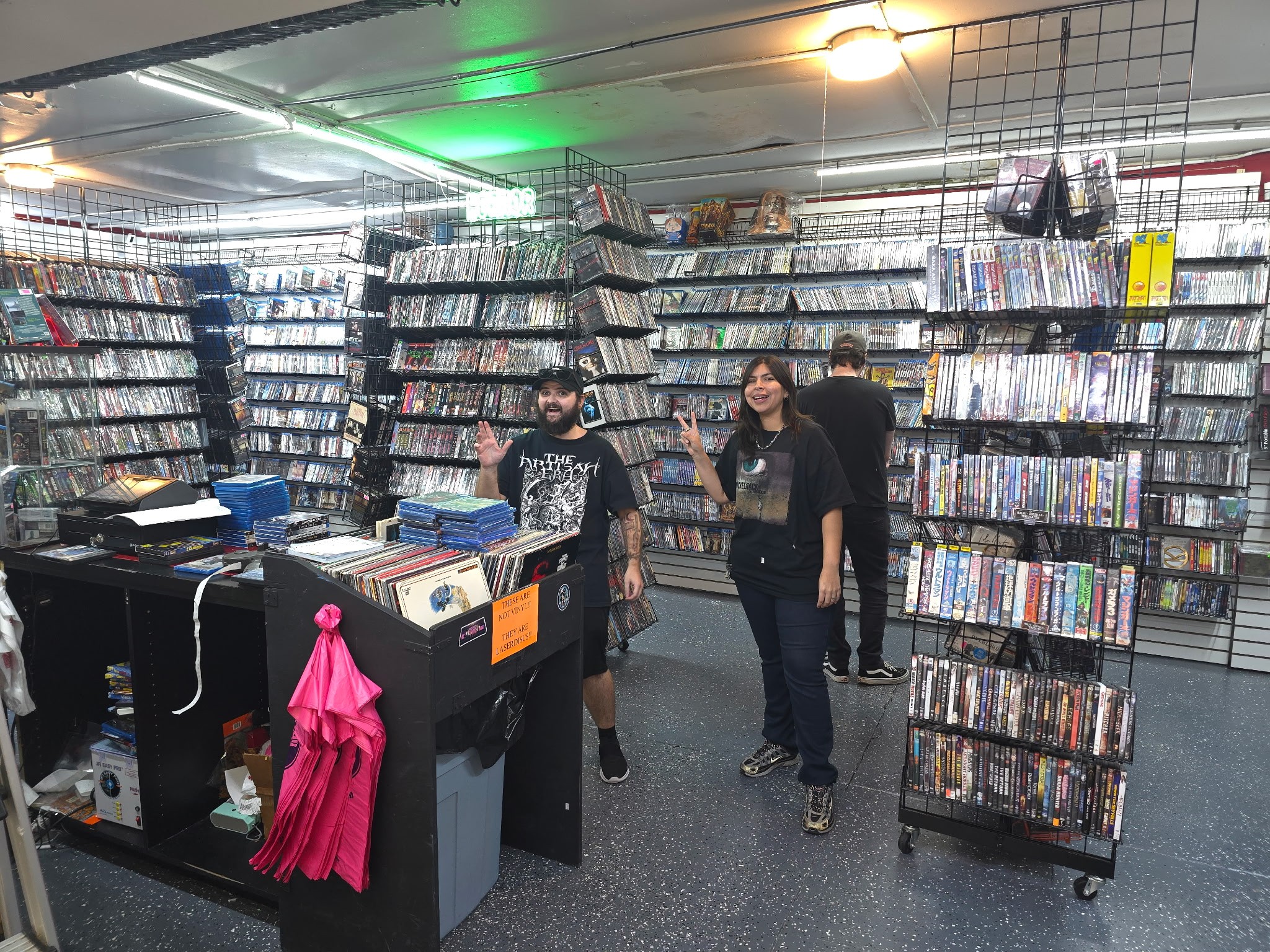 Three people browsing in a store with shelves filled with DVDs or video games, one person waving, store interior visible.