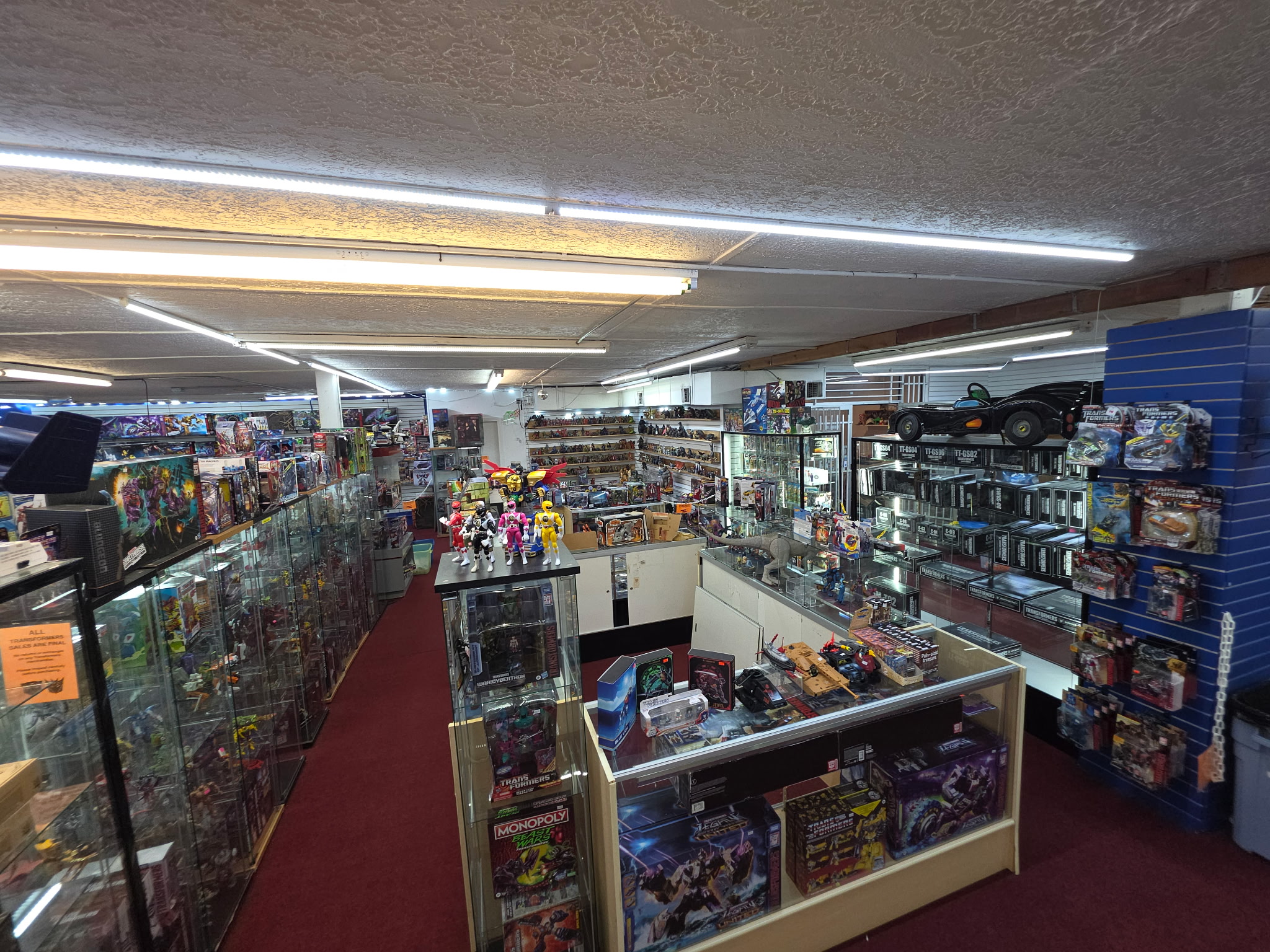 View of a store aisle with shelves stocked with toys, games, and merchandise, illuminated by fluorescent lights.