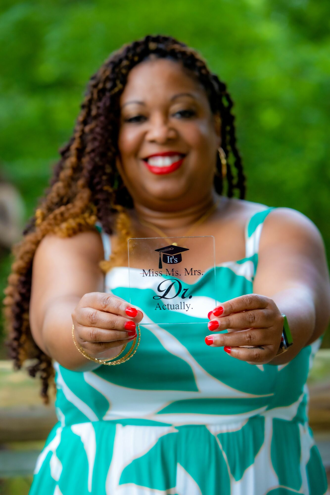 Woman smiling outdoors holding a glass with a message, wearing a teal and white dress, with green foliage background.