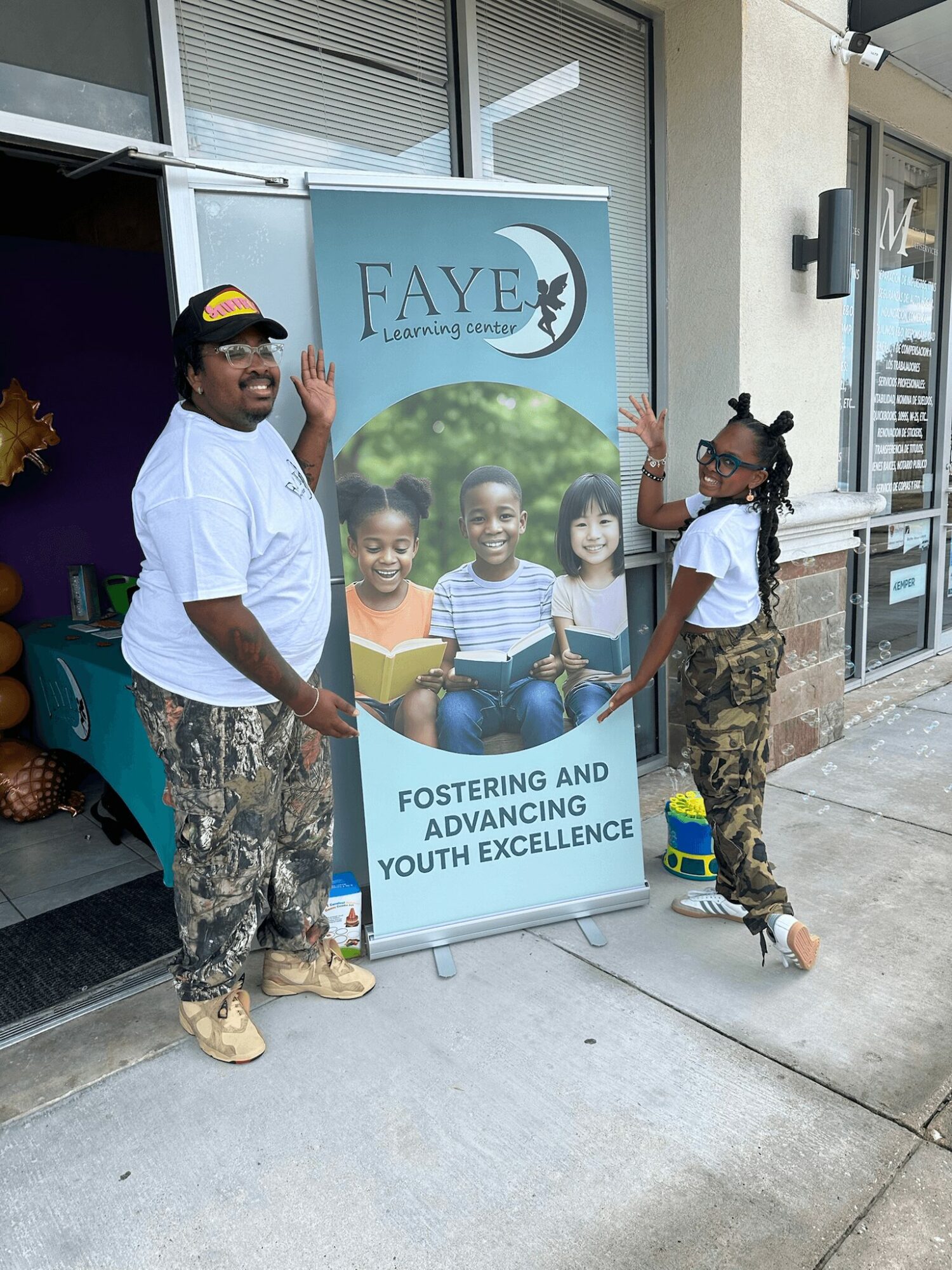 Two children and a woman stand outside a building next to a FAYE banner with children reading, smiling, and waving.