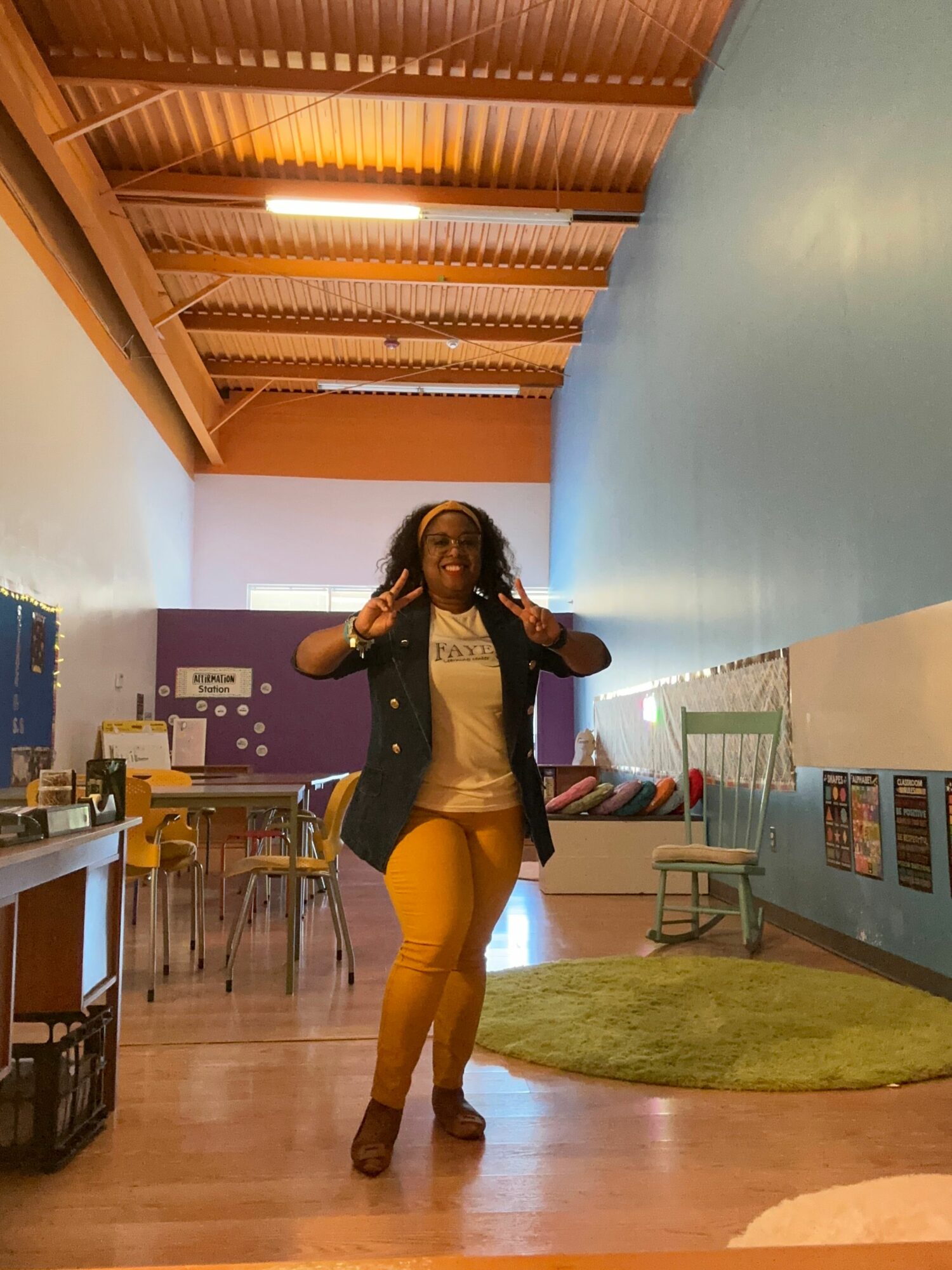 Woman standing in a room with wooden ceiling beams, a blue wall, and a green rug, holding a phone and smiling.
