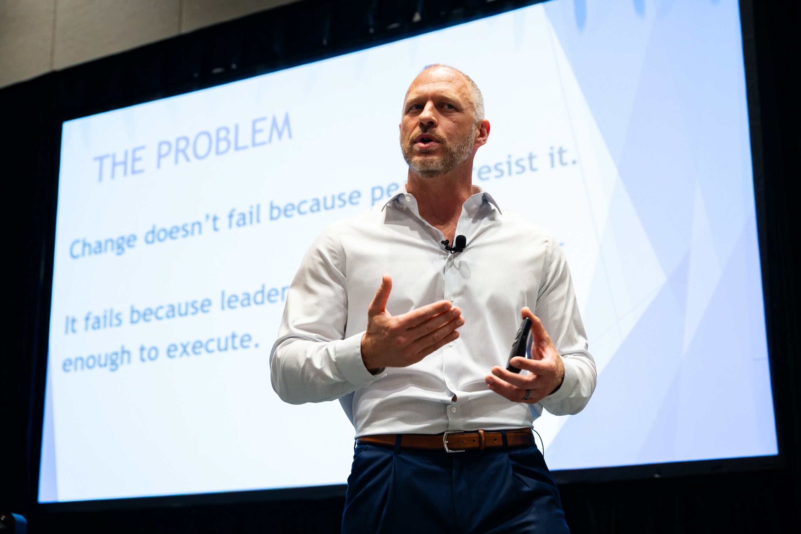 Man giving a presentation in front of a screen with text about the problem, gesturing with his right hand, wearing a white shirt and dark pants.