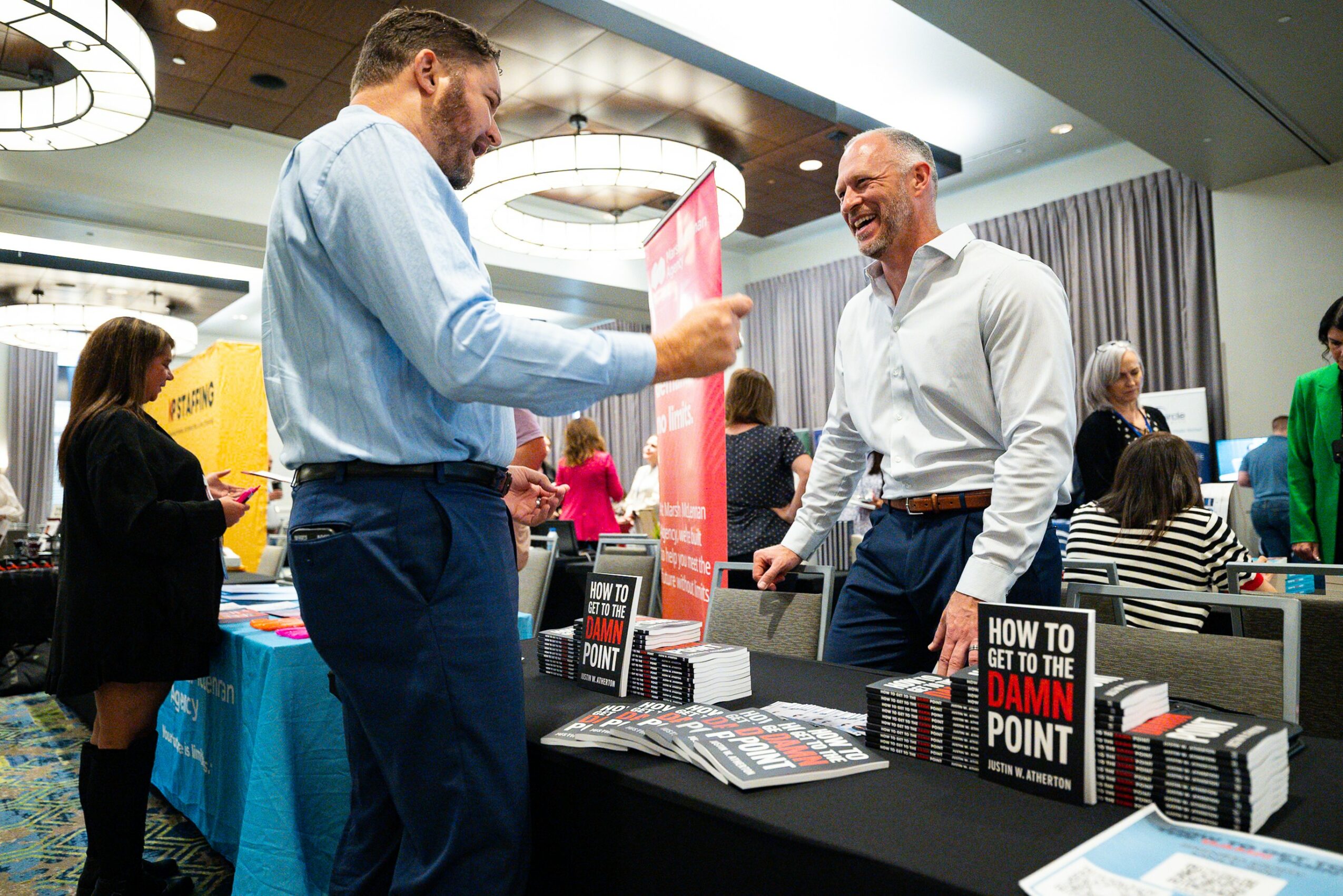 Two men smiling and talking at a booth with books and pamphlets, in a conference or event setting.