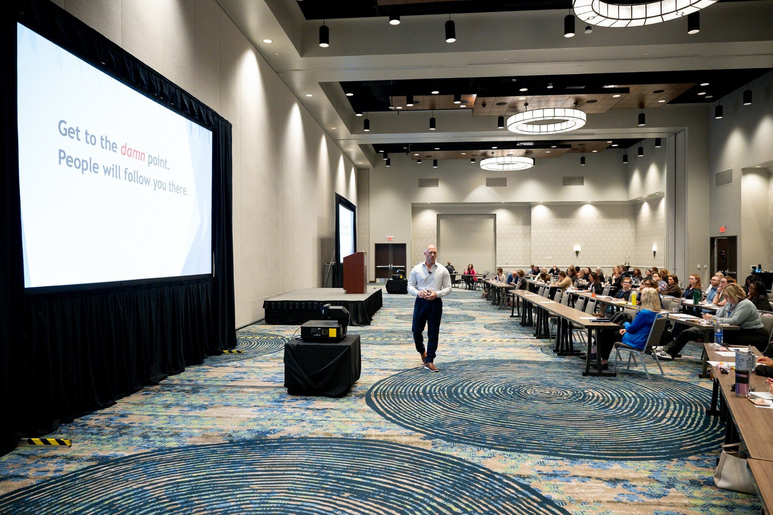 Conference room with a speaker walking towards a large screen displaying a presentation, audience seated at tables, ceiling lights, and patterned carpet.