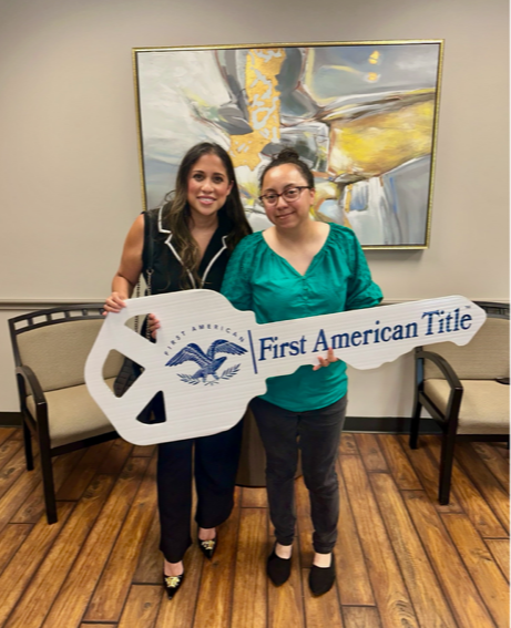 Two women standing indoors, holding a large key-shaped sign that reads 'First American Title,' with a painting behind them.