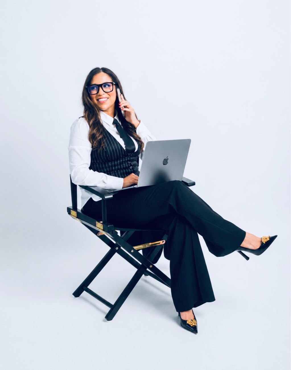 Woman sitting on a chair with a laptop, talking on a phone, smiling, wearing glasses and formal attire.