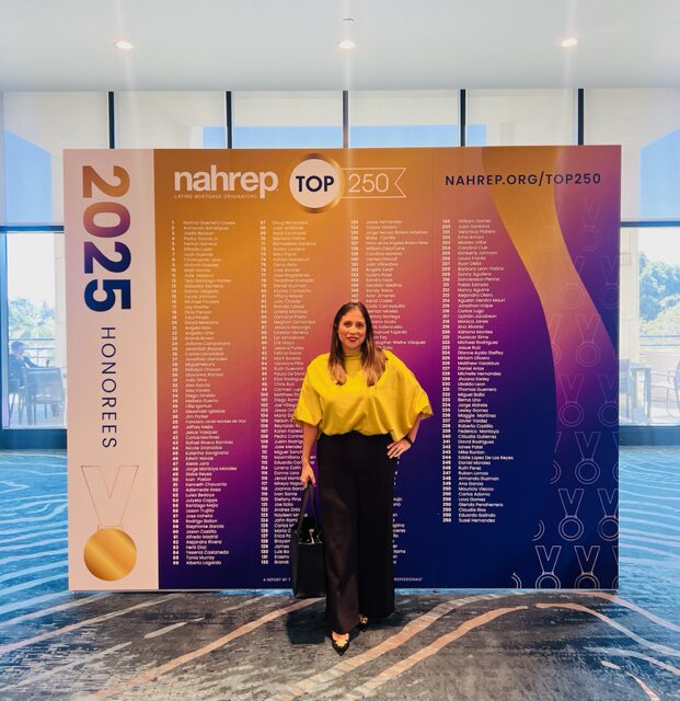 Woman in yellow top and black pants standing in front of a large event banner with text and logos, indoors with windows behind.
