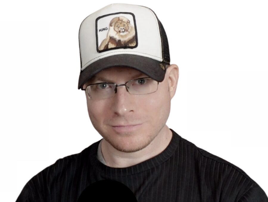Man wearing glasses and a trucker hat with a lion illustration, black shirt, neutral expression, white background.