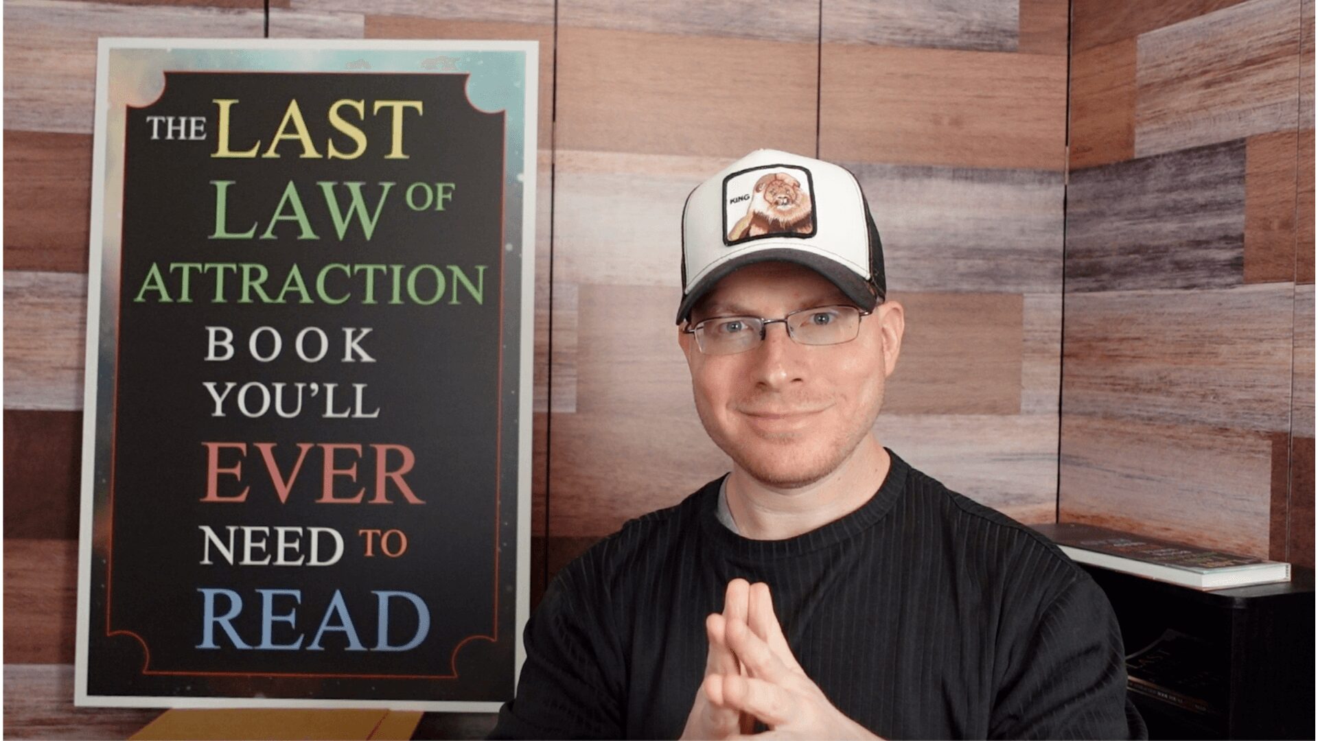 Man wearing glasses and a cap, sitting with hands clasped, in front of a wooden wall and a book sign.